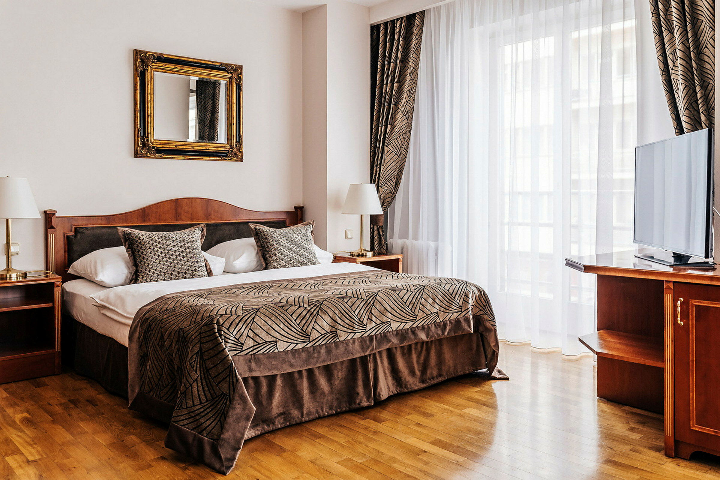 Ett elegant hotellrum med en stor säng, klädd i ett mönstrat överkast och prydnadskuddar. Rummet har en spegel ovanför sängen, två nattduksbord med lampor och en TV på en möbel bredvid en stor fönsteryta. (AI-genererad text)