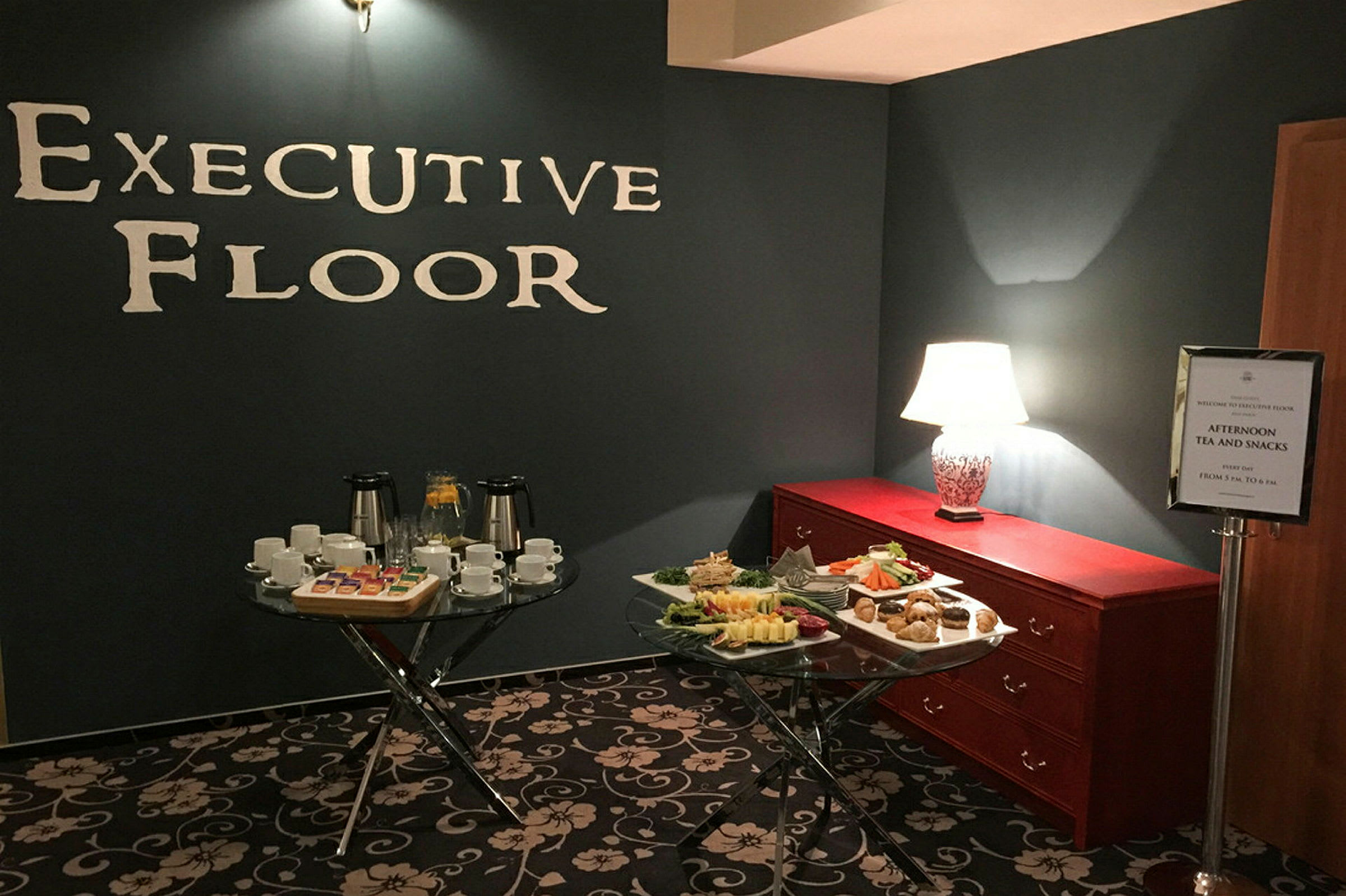 Ett rum med texten "Executive Floor" på väggen, en modern belysning, två bord med koppar, tallrikar och tilltugg samt en byrå med bordslampa. (AI-genererad text)