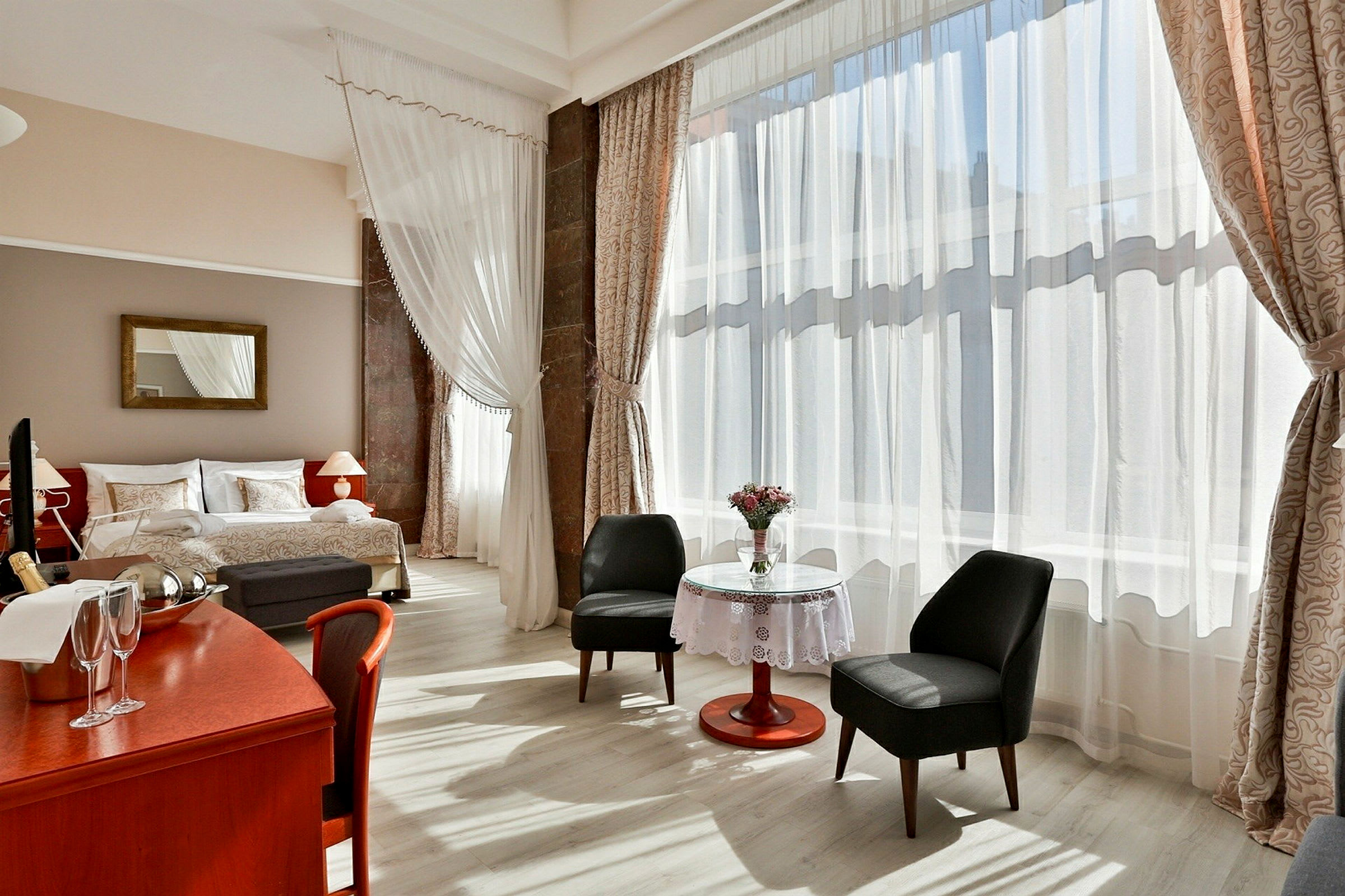 Ett hotellrum med stor fönsteryta och mycket dagsljus. Rummet har en dubbelsäng, en läshörna med två stolar och ett bord med vas och blommor, samt ett skrivbord med champagneflaska och glas. (AI-genererad text)
