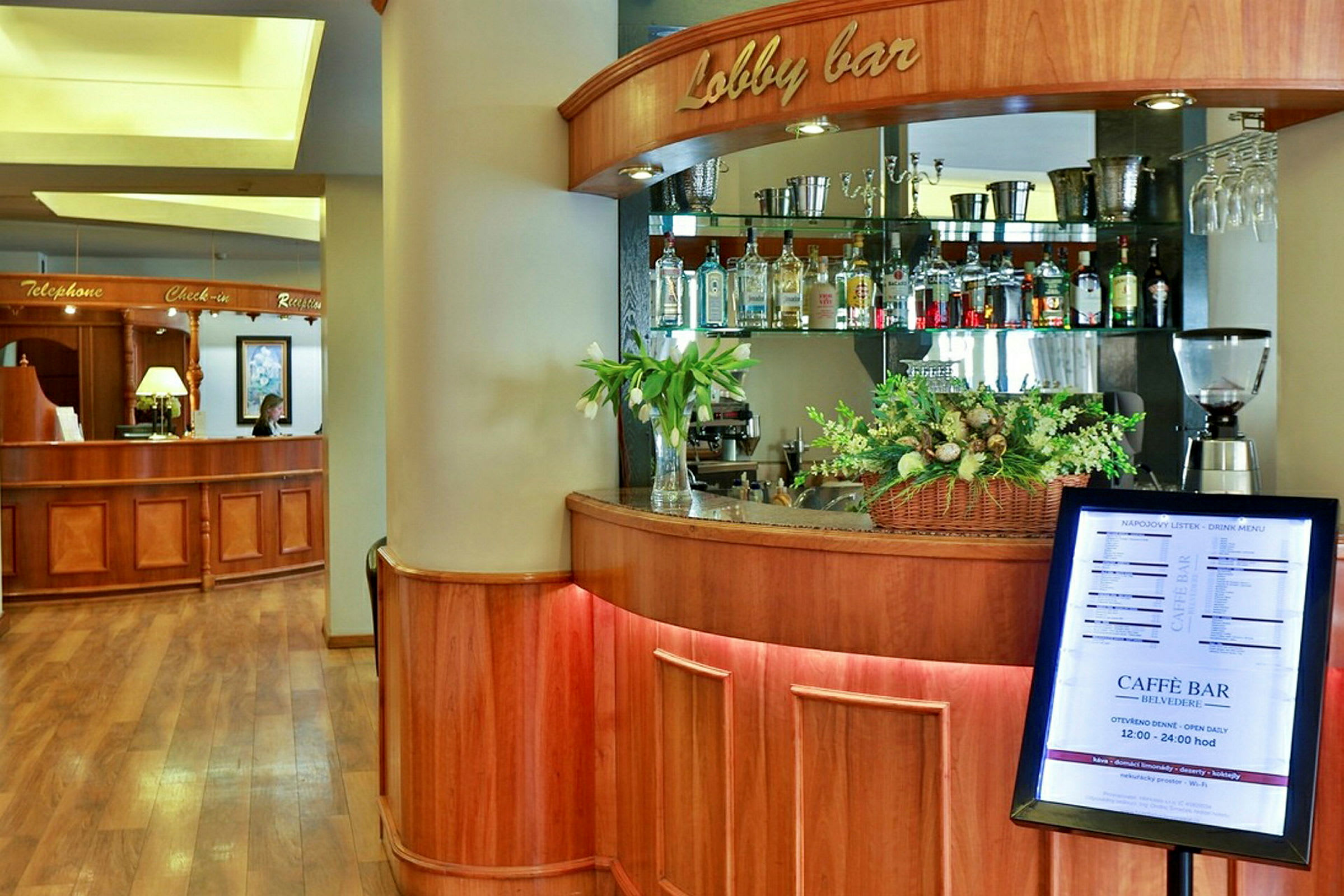 En hotellobbymiljö med en receptionsdisk i trä på vänster sida och en lobbybar med dryckesflaskor och blomdekorationer på höger sida. (AI-genererad text)