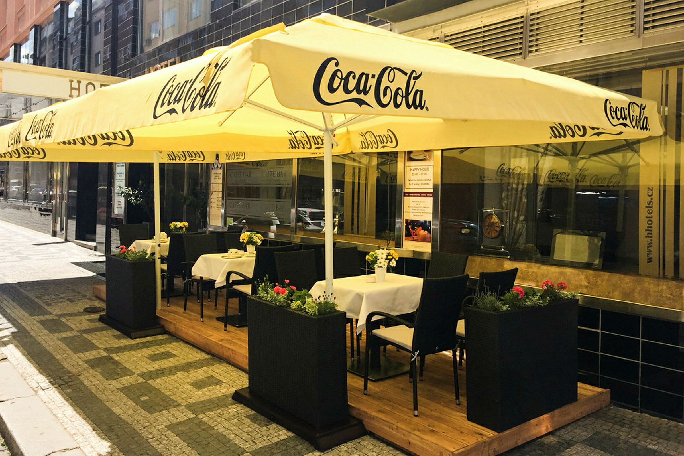 Ett utecafé med bord och stolar under stora gula parasoller märkta med Coca-Cola-logotypen. (AI-genererad text)