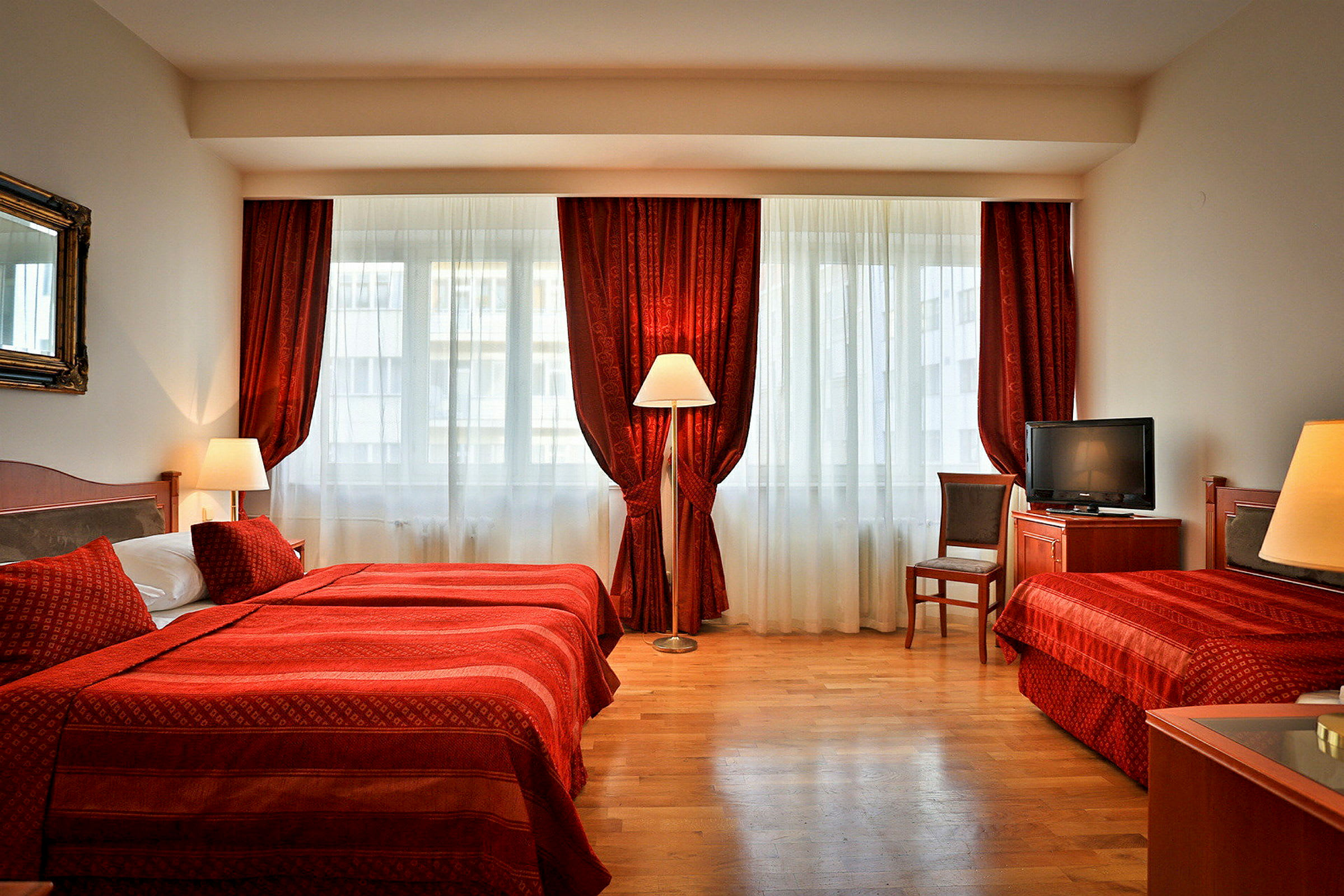 Ett hotellrum med två sängar, röda sängkläder och gardiner samt en TV och en stol. (AI-genererad text)