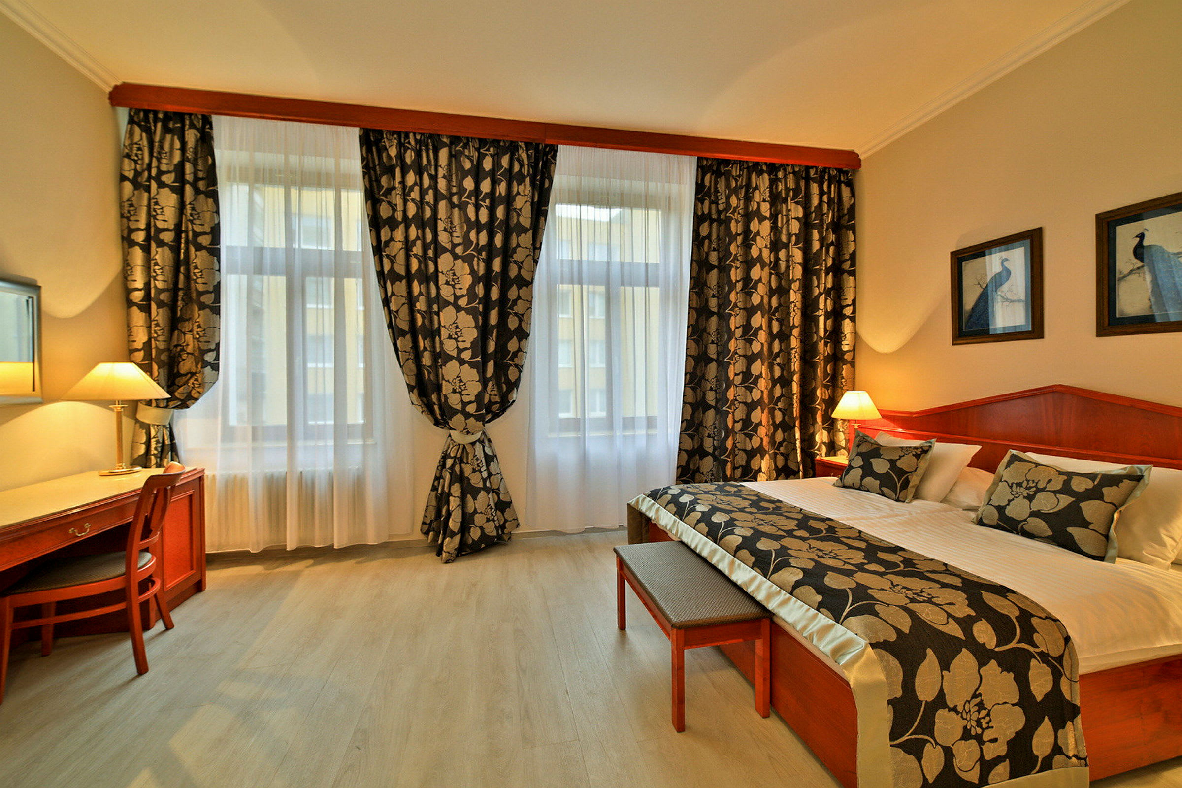 Ett hotellrum med två stora fönster, en dubbelsäng med dekorativa kuddar och överkast, samt ett skrivbord med stol och lampa. (AI-genererad text)
