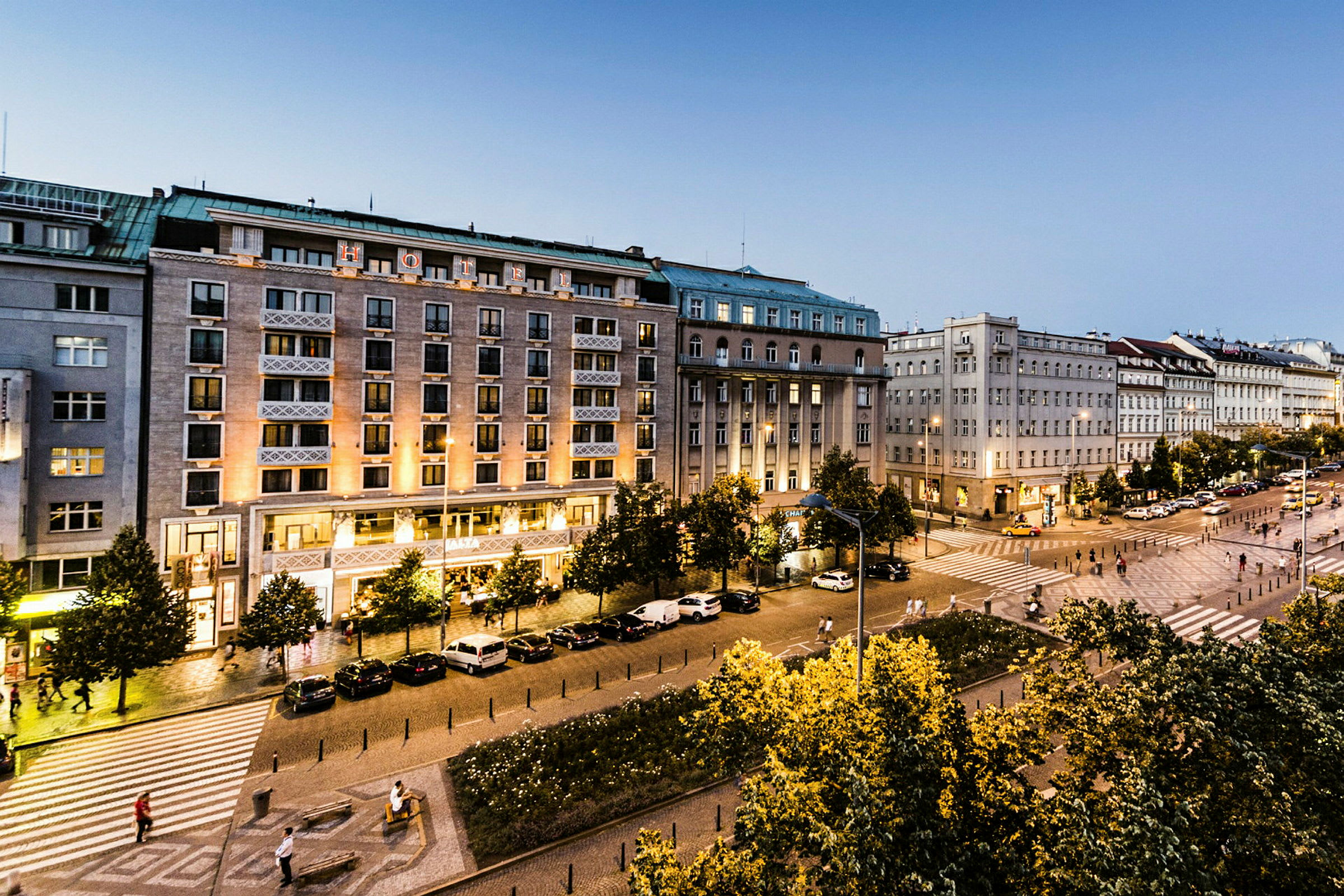 Bild av en stadsgata med flera byggnader, träd och parkerade bilar. Gatubelysning lyser upp området och fotgängare rör sig längs trottoaren. (AI-genererad text)