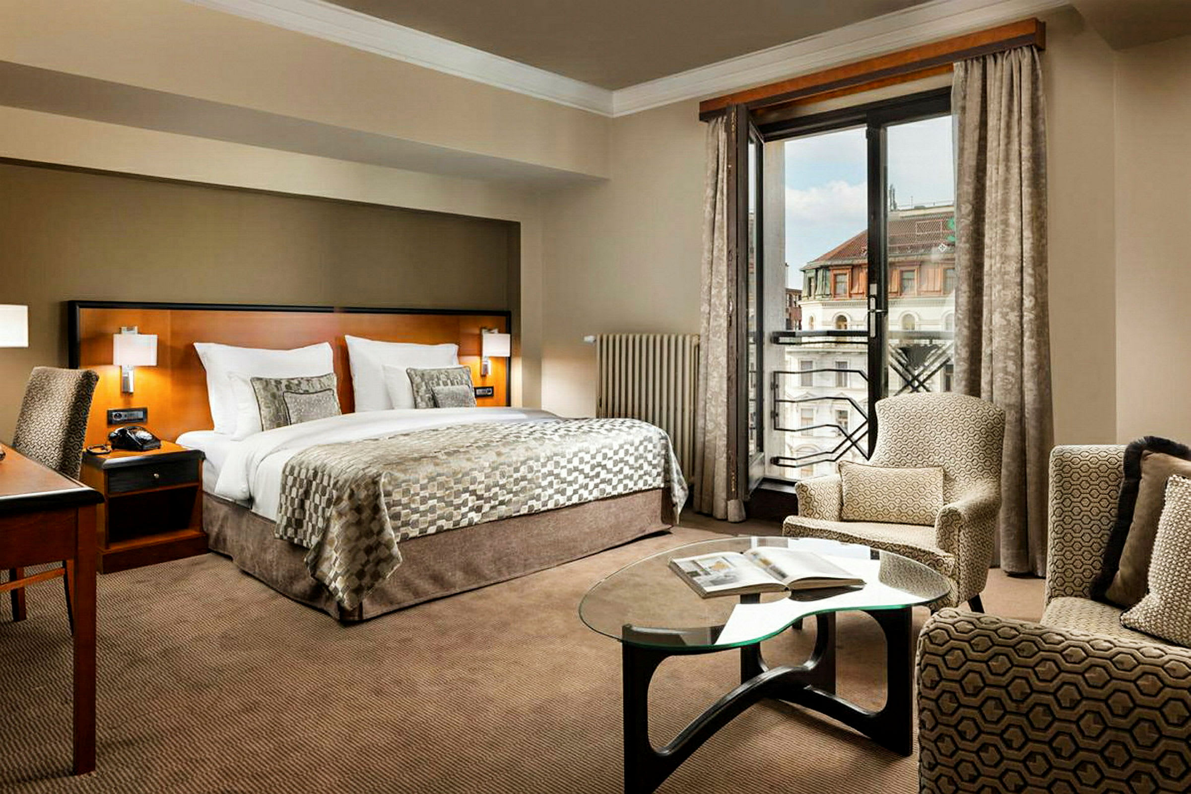 Ett elegant hotellrum med en dubbelsäng, en arbetsyta, en sittgrupp och ett fönster som visar stadsvy. (AI-genererad text)