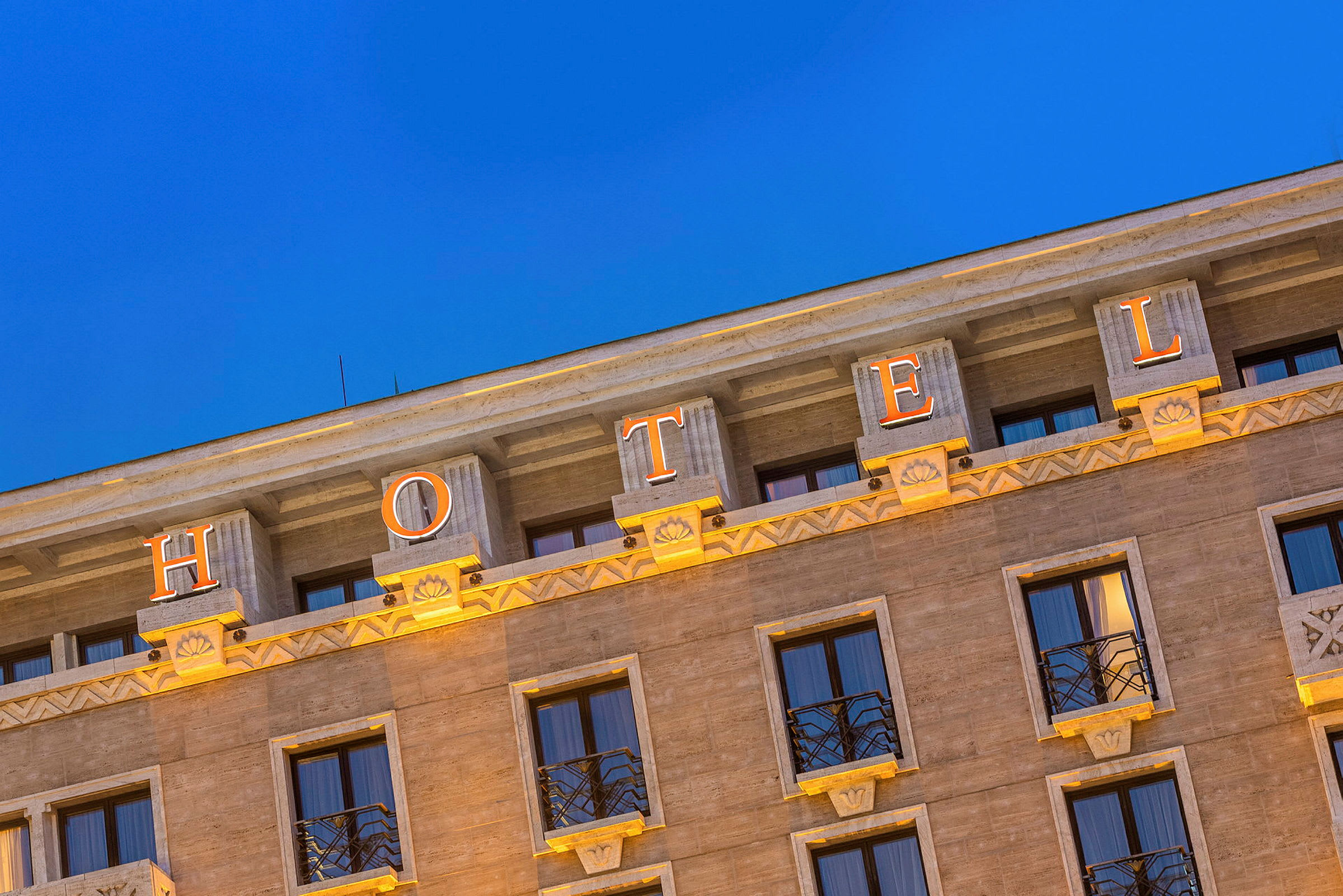Fasaden av ett hotell med upplysta bokstäver som stavar ordet 'HOTEL' mot en klarblå kvällshimmel. (AI-genererad text)