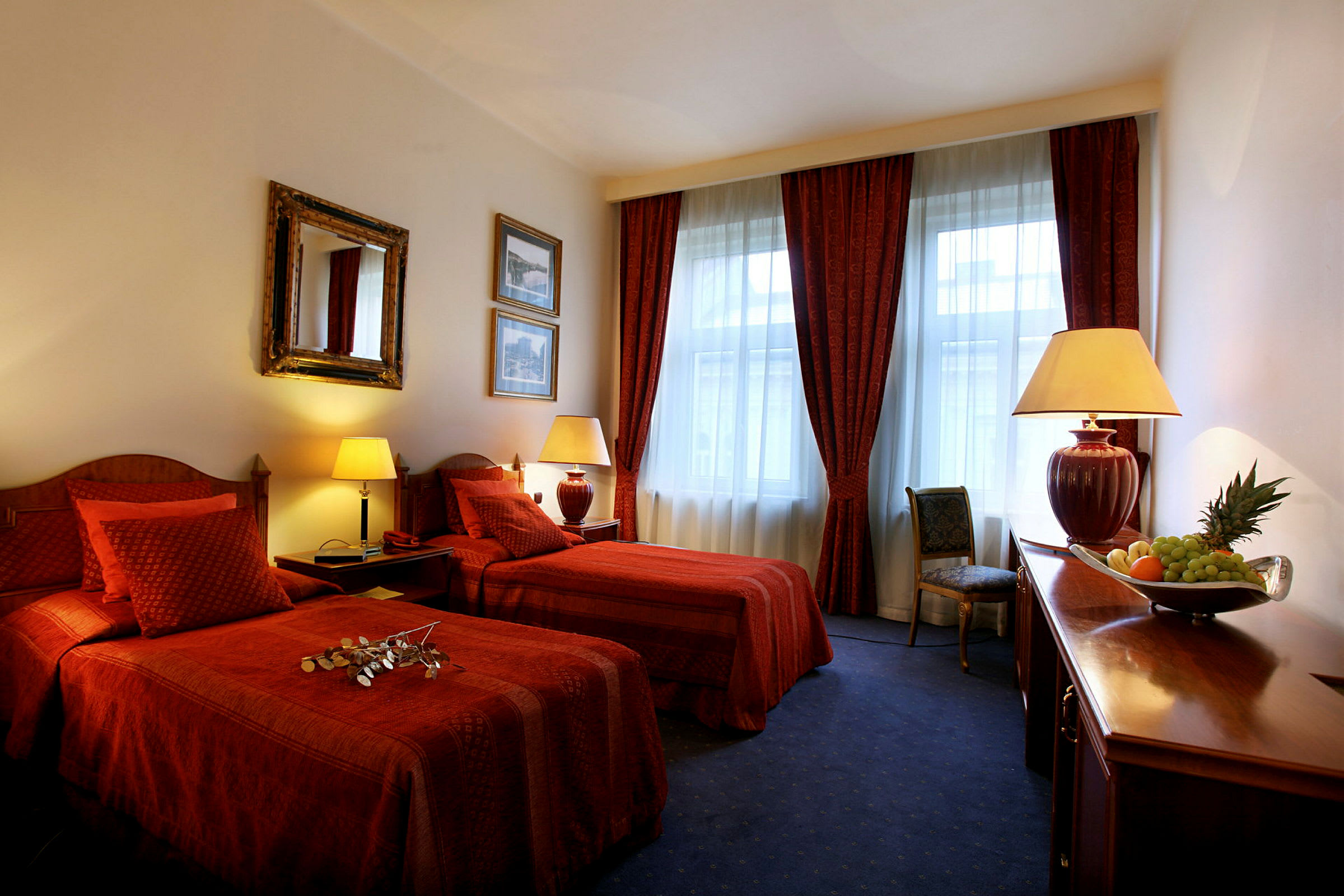 Ett hotellrum med två enkelsängar, röd inredning och stora fönster med gardiner. Ett bord med en skål frukt är också synligt. (AI-genererad text)