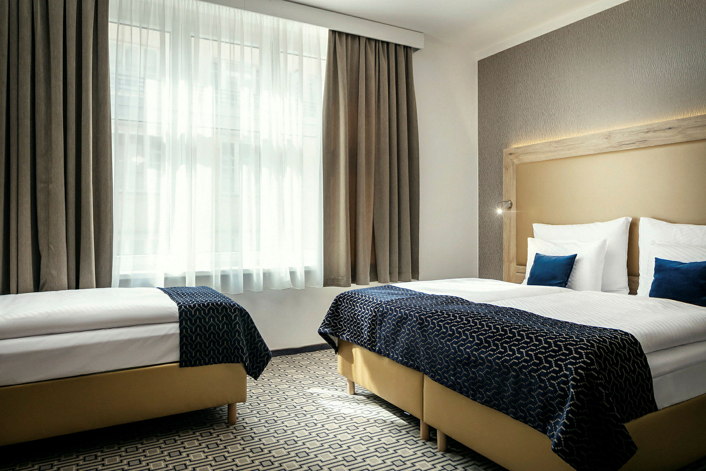 Ett hotellrum med två enkelsängar, vita lakan, blåa dekorationskuddar och ett stort fönster med vita gardiner. (AI-genererad text)