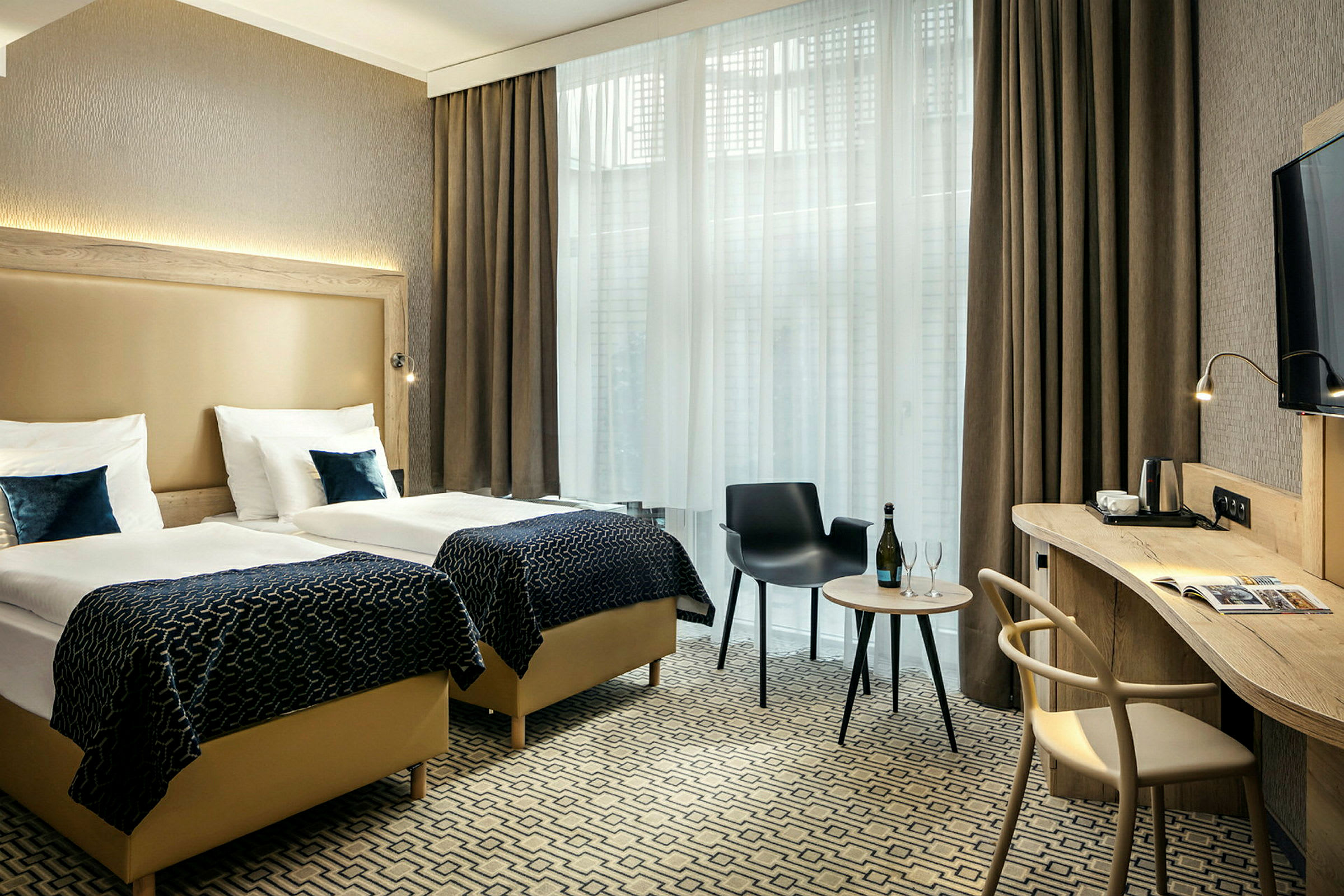 Hotellrum med två enkelsängar, skrivbord, stol och ett litet bord med flaska samt glas framför stora fönster med gardiner. (AI-genererad text)