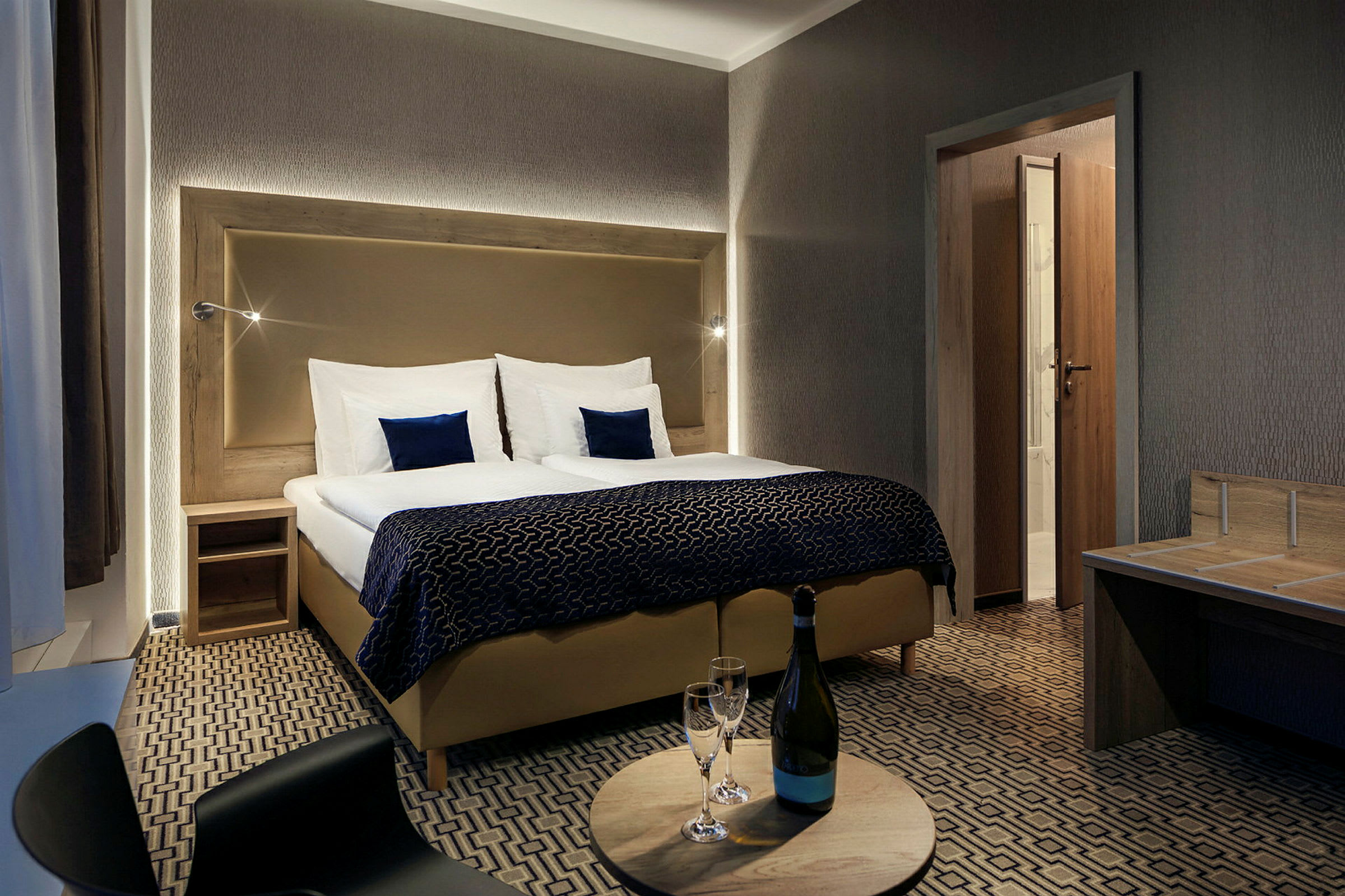 Ett hotellrum med dubbelsäng, sängbord, och en rund bord med en flaska och två glas. (AI-genererad text)