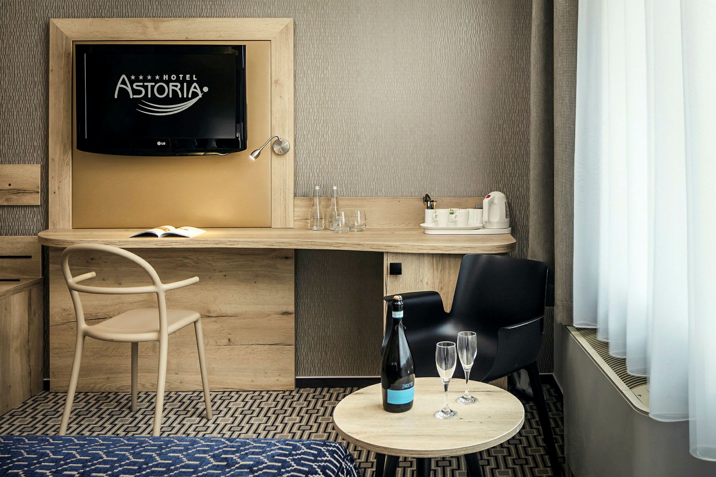 Ett hotellrum med en TV på väggen, ett skrivbord med en stol, en vattenkokare och glas, samt ett runt bord med en flaska och två glas. (AI-genererad text)