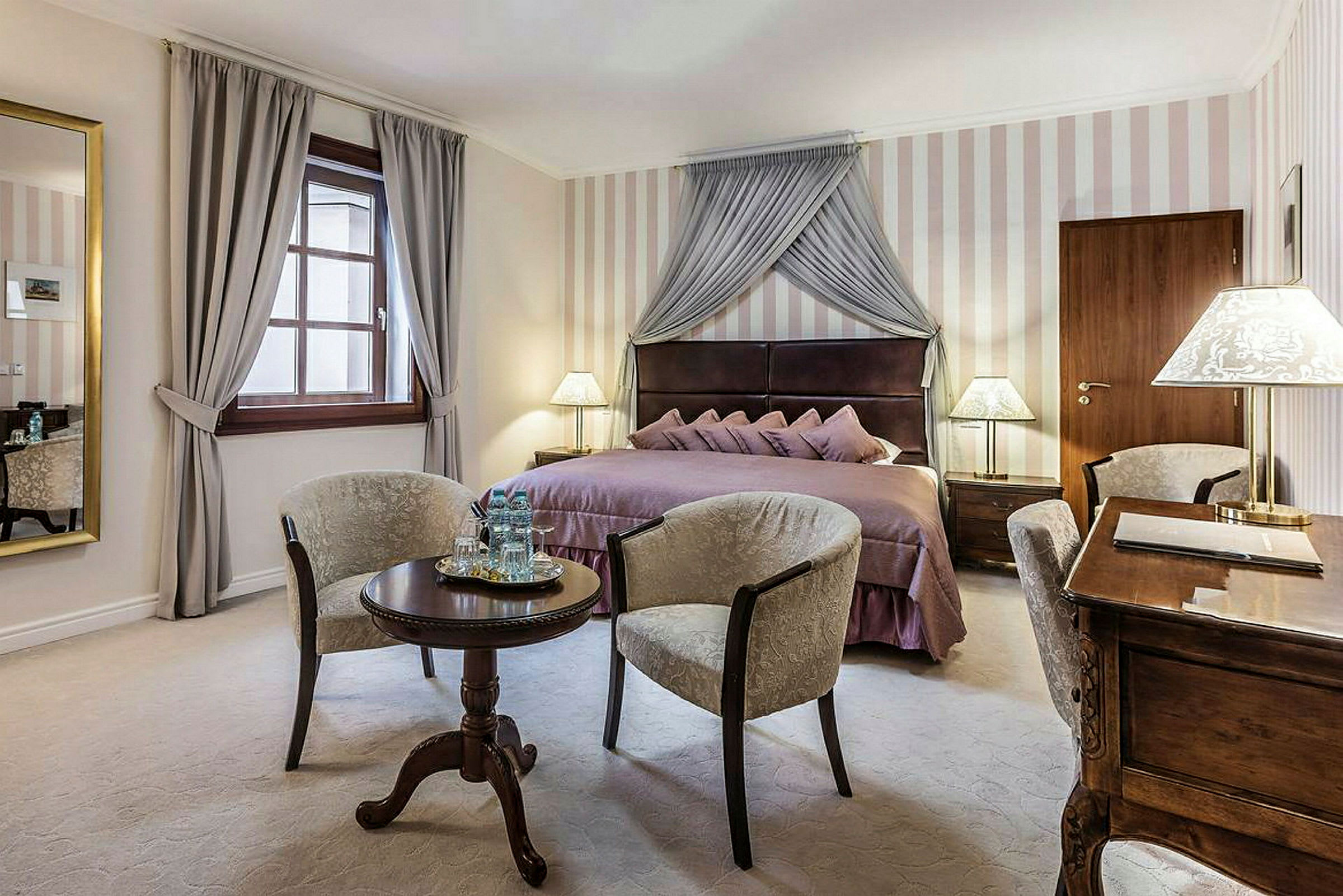 Ett elegant hotellrum med en dubbelsäng klädd i lila sängkläder, två fåtöljer placerade runt ett litet runt bord, och ett skrivbord med lampa. (AI-genererad text)