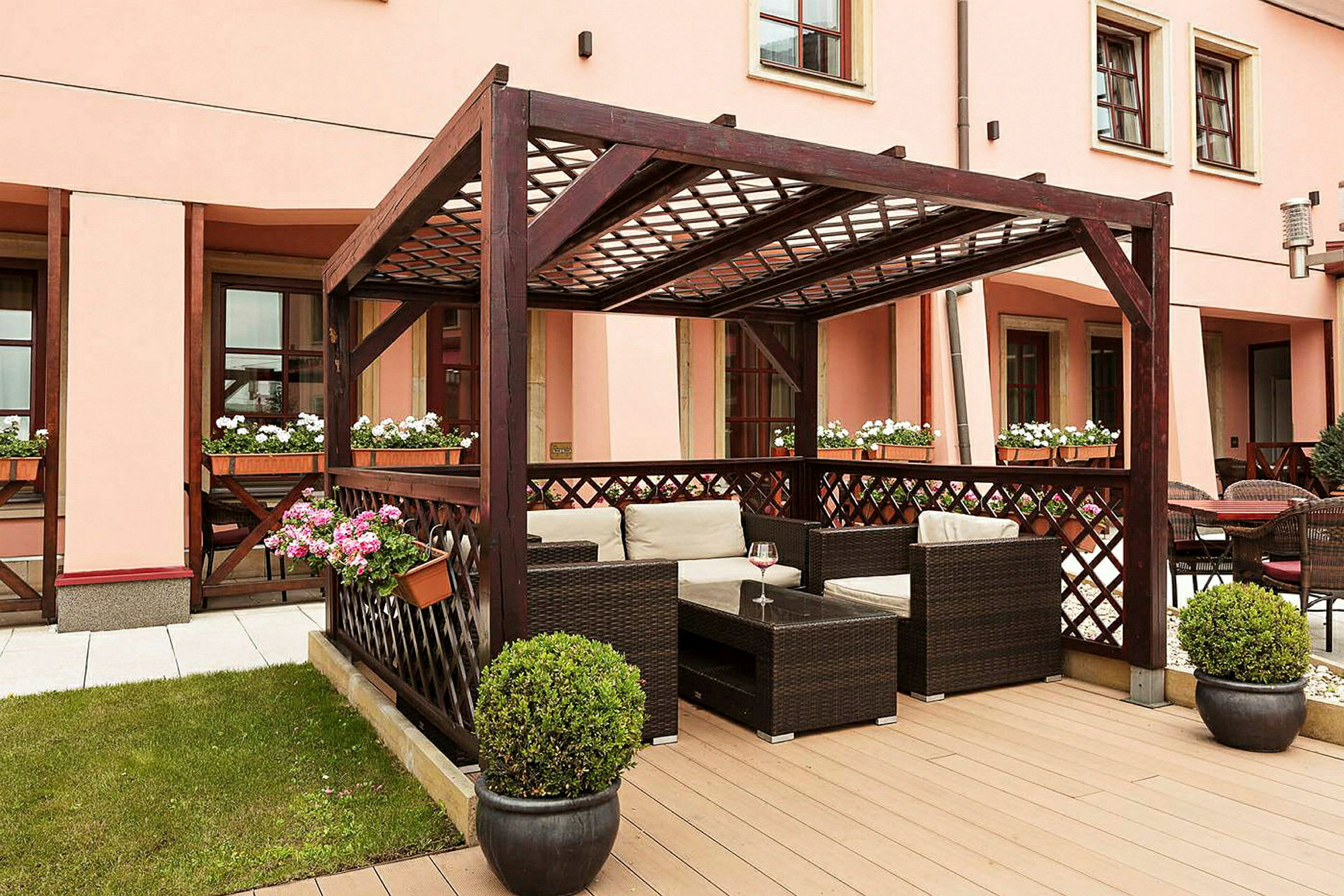 Bilden visar en uteplats med en pergola i mörkt trä, möblerade med rottingstolar och bord. Blomkrukor med färgglada blommor pryder området. (AI-genererad text)