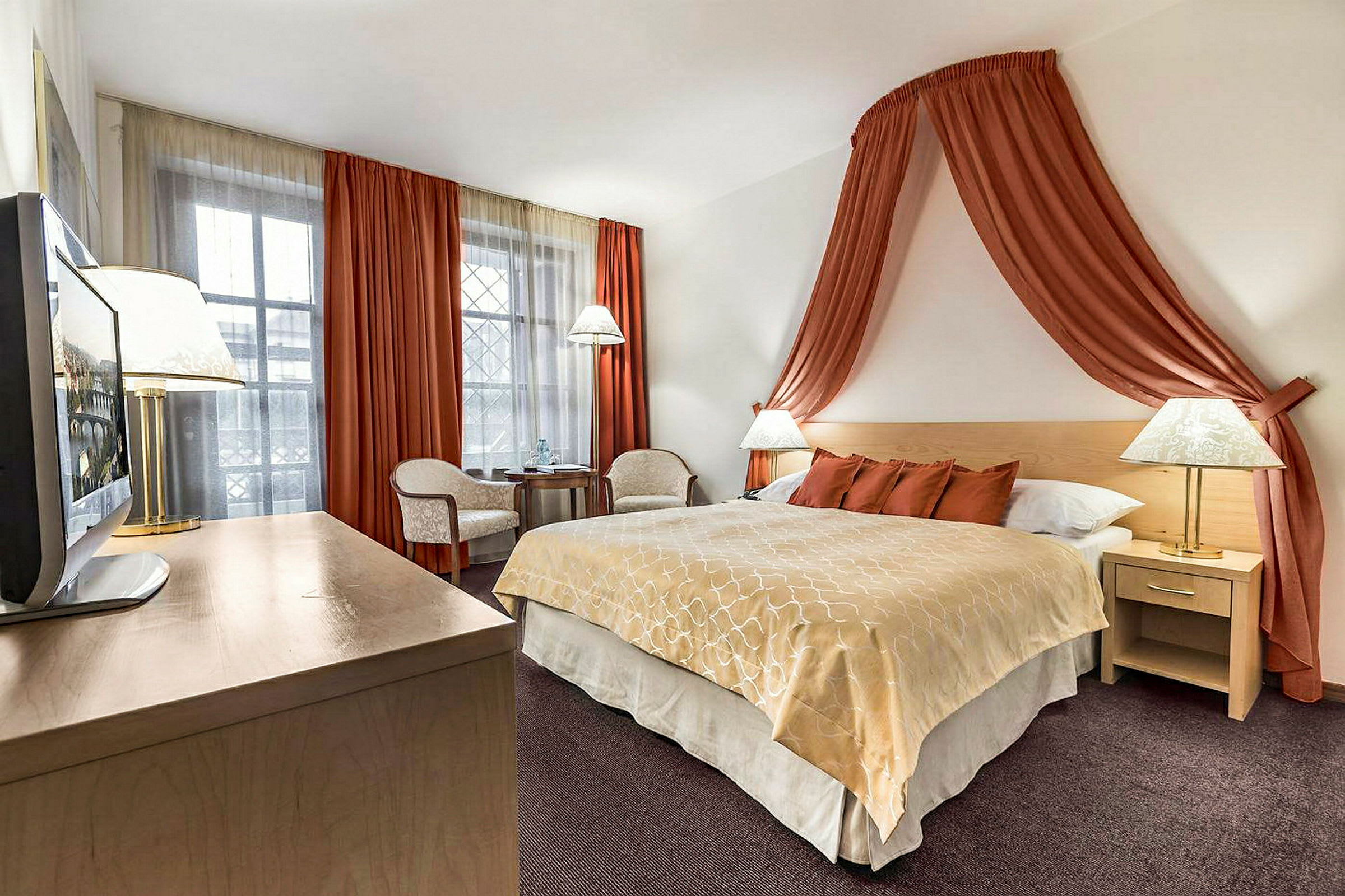 Ett hotellrum med en stor säng täckt med beige och orange sängkläder. Sängen är omgiven av nattduksbord med lampor och har orange gardiner hängande ovanför som draperi. I hörnet finns ett litet bord med två stolar framför ett fönster täckt med vita och orange gardiner. (AI-genererad text)