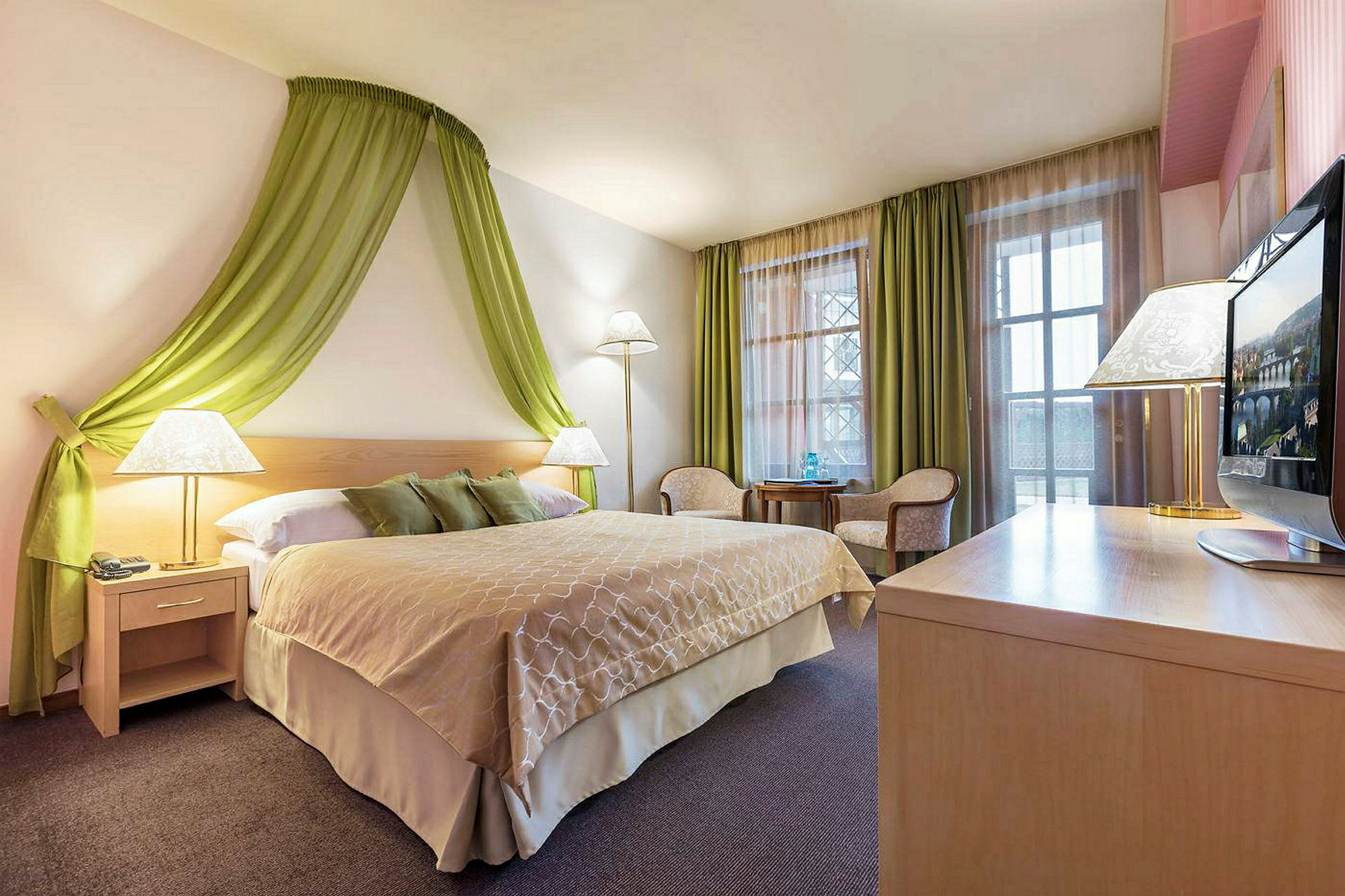 Ett hotellrum med en dubbelsäng, gröna gardiner och väggdekorationer, sittmöbler och ett skrivbord med en TV. (AI-genererad text)