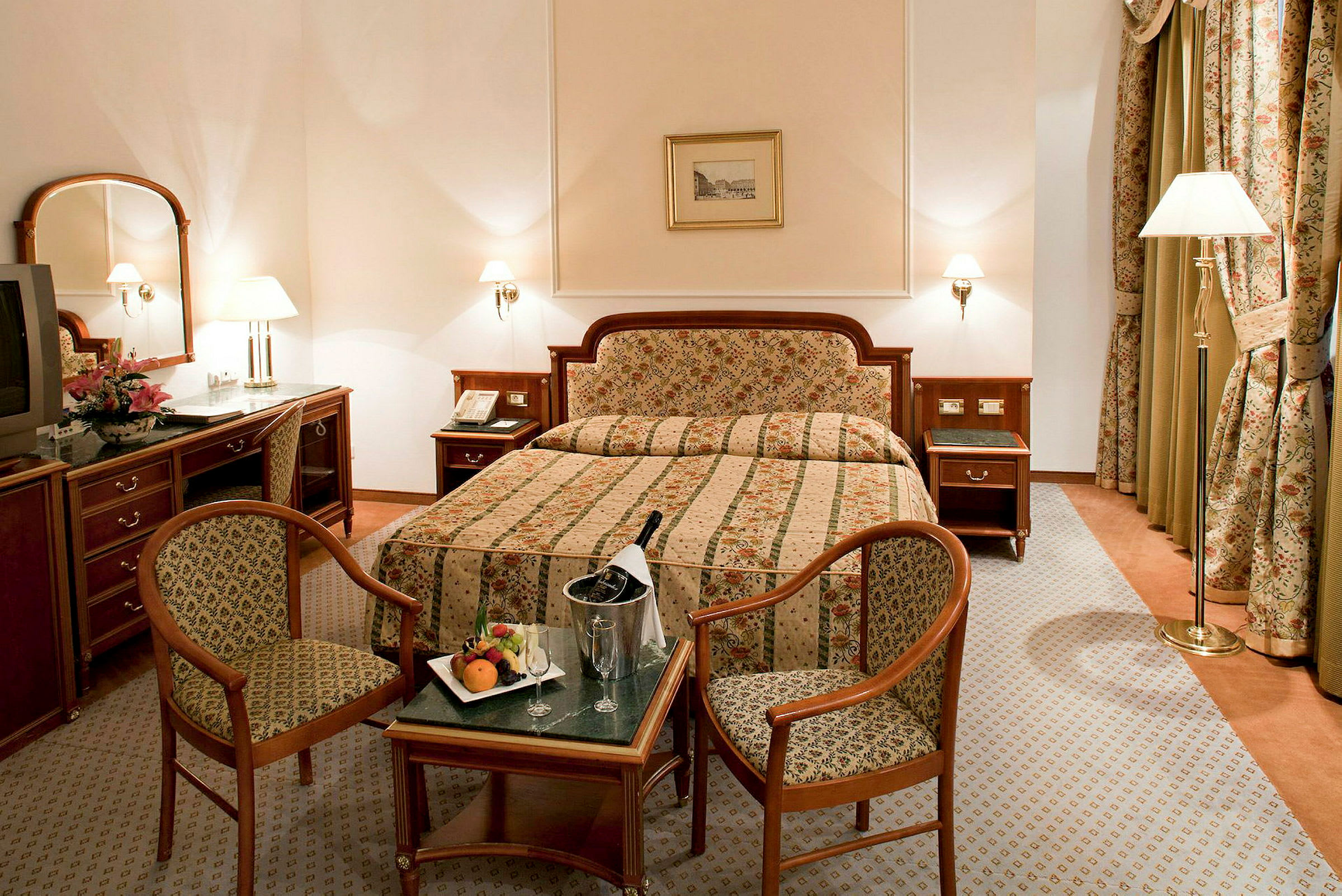 Hotellrum med dubbelsäng, två stolar, bord med frukt och champagne samt klassisk inredning. (AI-genererad text)