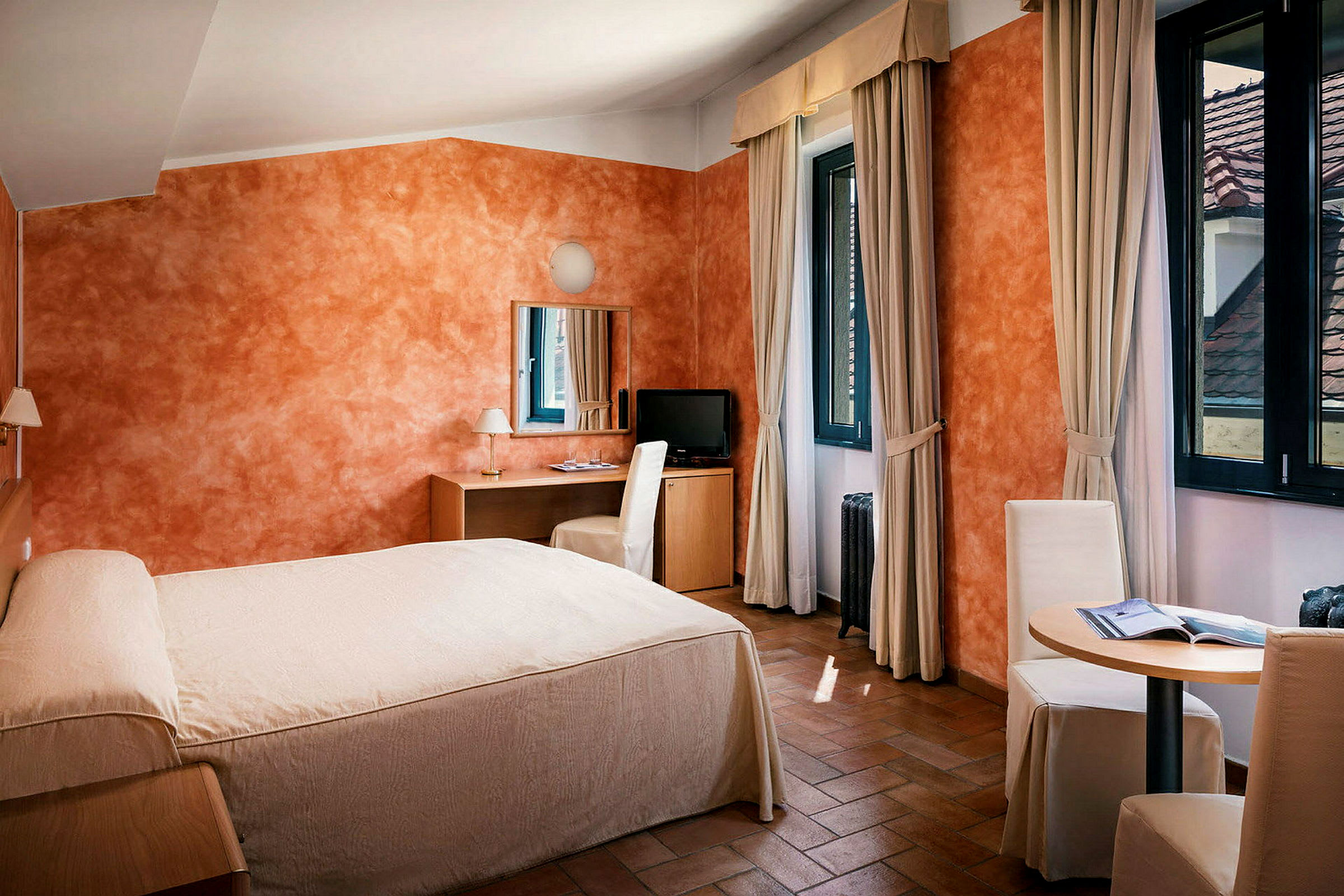 Ett hotellrum med säng, skrivbord och stol, samt ett litet bord med två stolar. Rummet har orange strukturerade väggar och fönster med gardiner. (AI-genererad text)