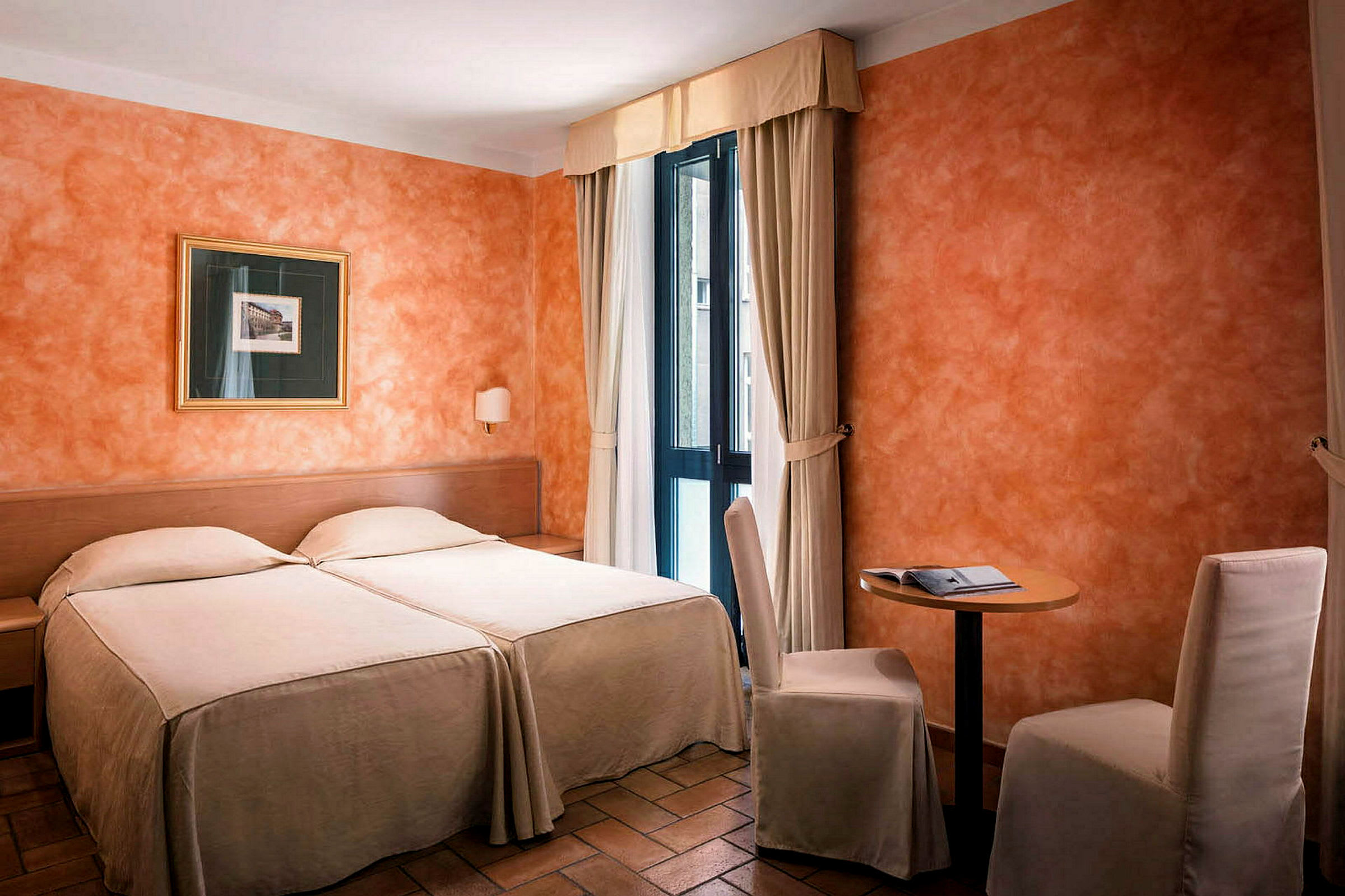 Ett hotellrum på bilden med bäddar i ljusa överdrag, orange väggar, en liten bord med två stolar och en tavla. Fönster med ljusa gardiner släpper in dagsljus. (AI-genererad text)