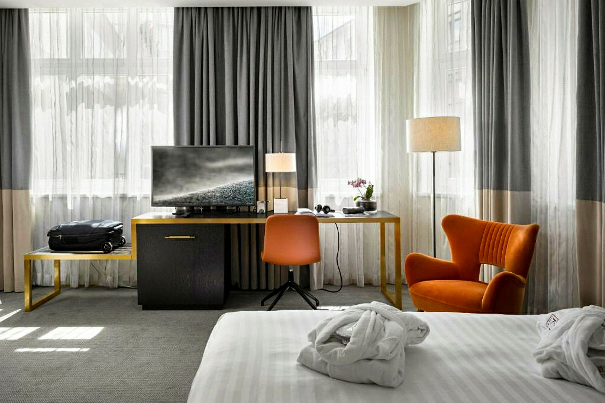 Ett hotellrum med en säng med en vit sängklädsel i förgrunden, och en sittgrupp med orange stol och skrivbord med en TV i bakgrunden. (AI-genererad text)