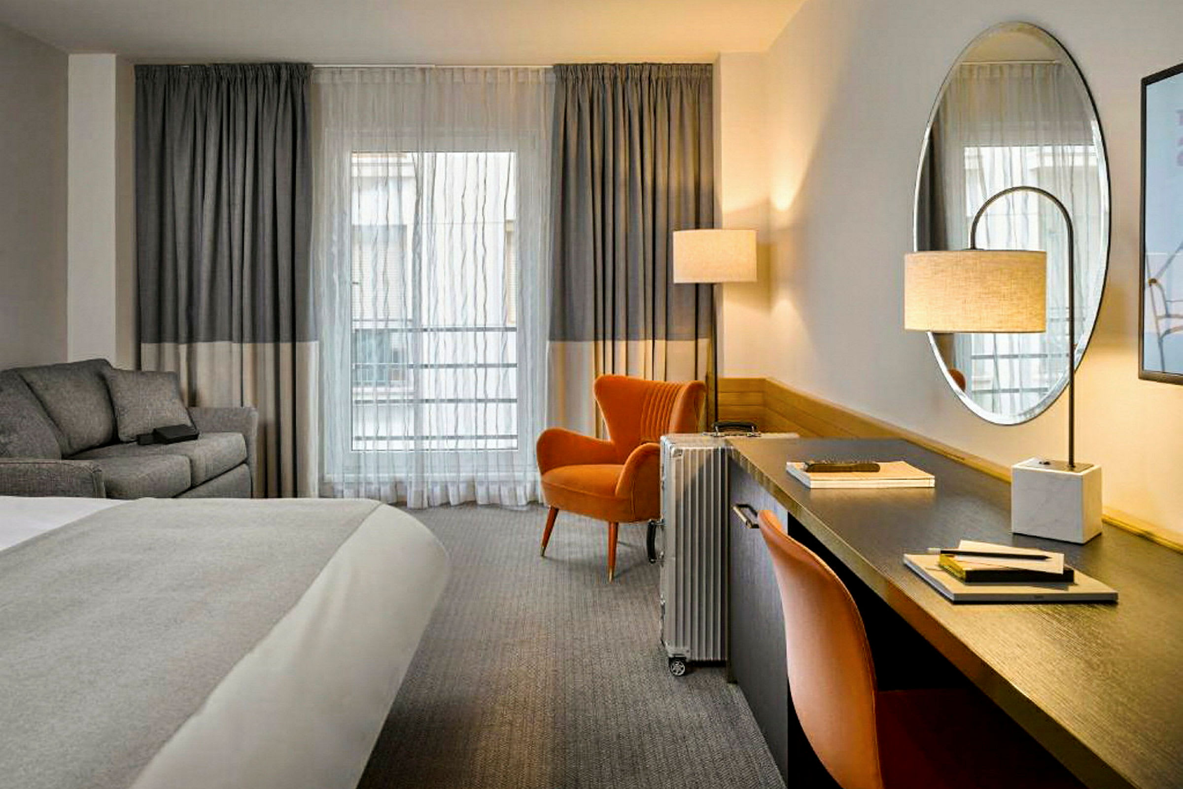 Ett hotellrum med en säng, skrivbord, och en grå soffa. Fönstret har grå gardiner och en orange fåtölj med en golvlampa bredvid. (AI-genererad text)