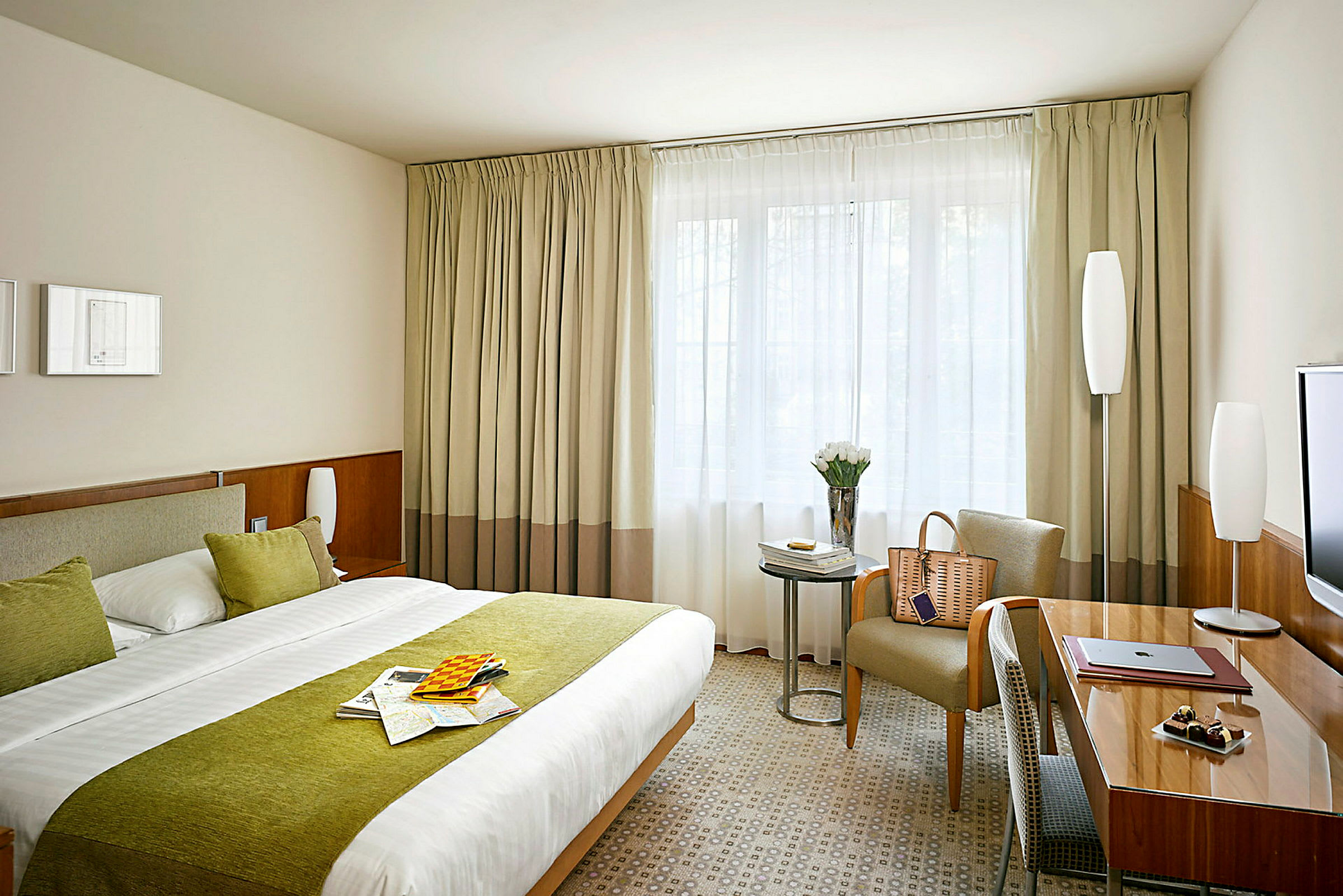 Hotellrum med dubbelsäng, gröna detaljer, skrivbord, stol och fönster med gardiner. (AI-genererad text)