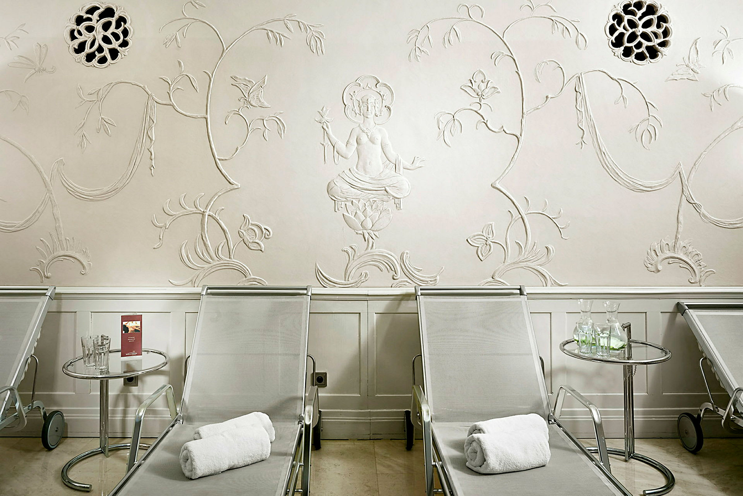 Tre solstolar på rad framför en ljus vägg med dekorationer, inklusive en relief av en sittande figur. (AI-genererad text)