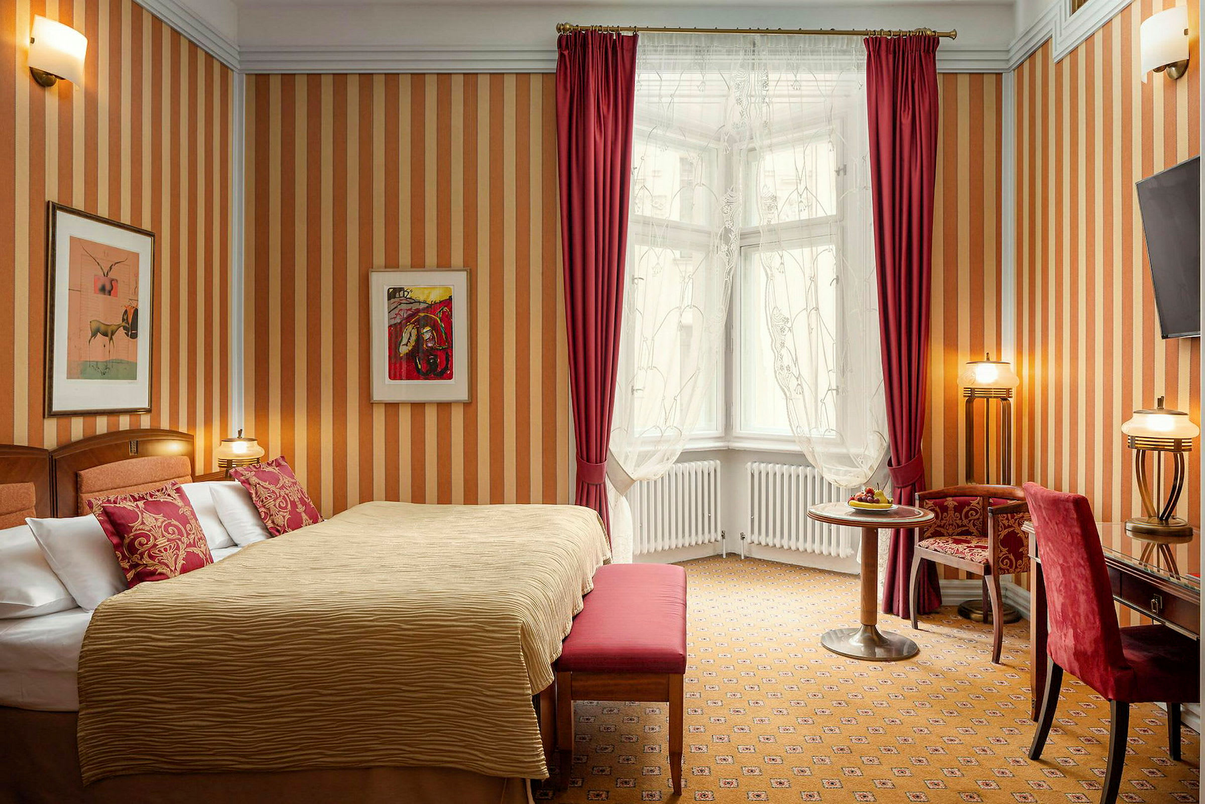 Ett hotellrum med en bred säng, röda kuddar och gardiner, samt en liten sittgrupp framför ett fönster. (AI-genererad text)