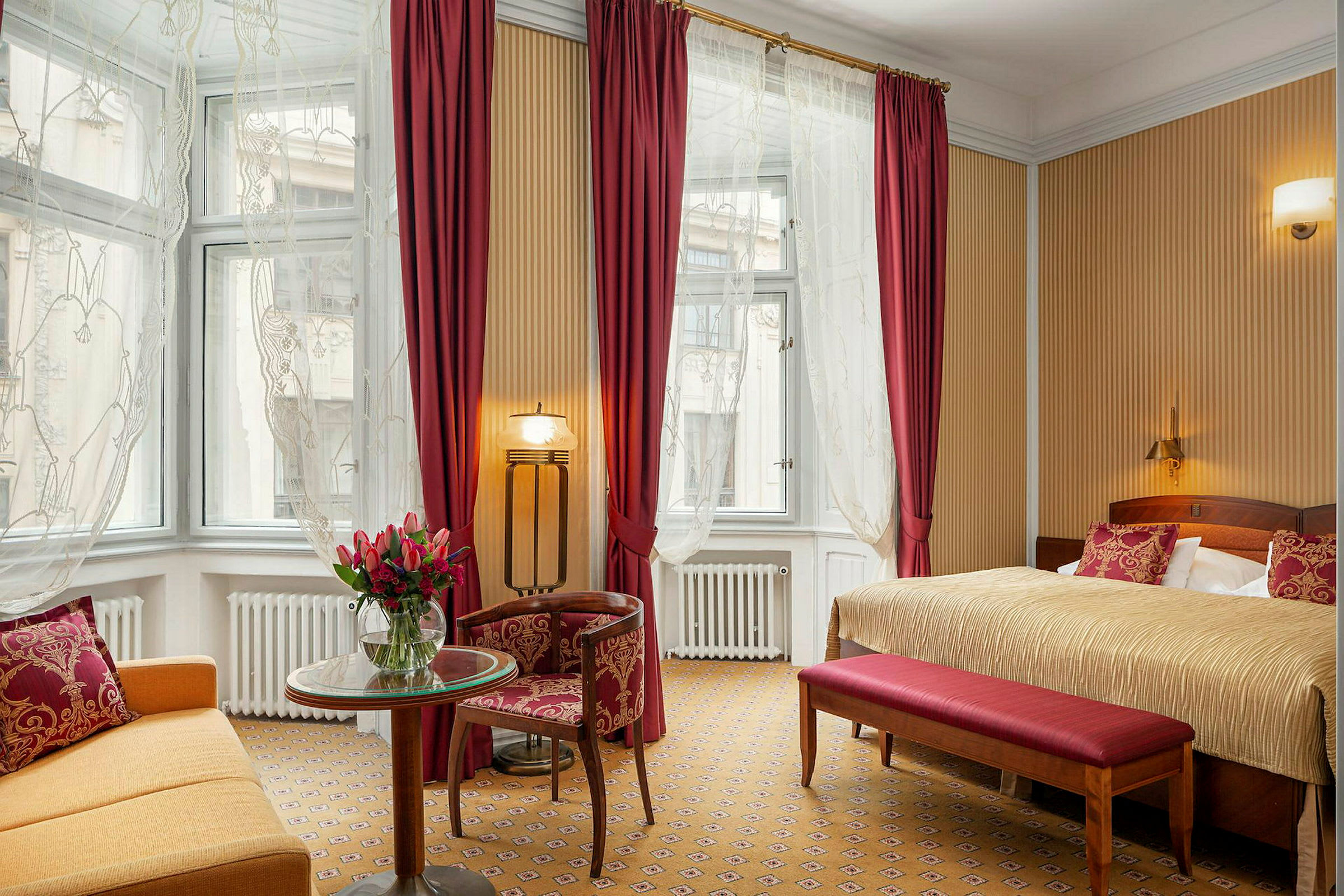 Ett hotellrum med en stor säng, sittplats, och bord. Rummet har en varm inredning med röda gardiner och dekorationer. (AI-genererad text)