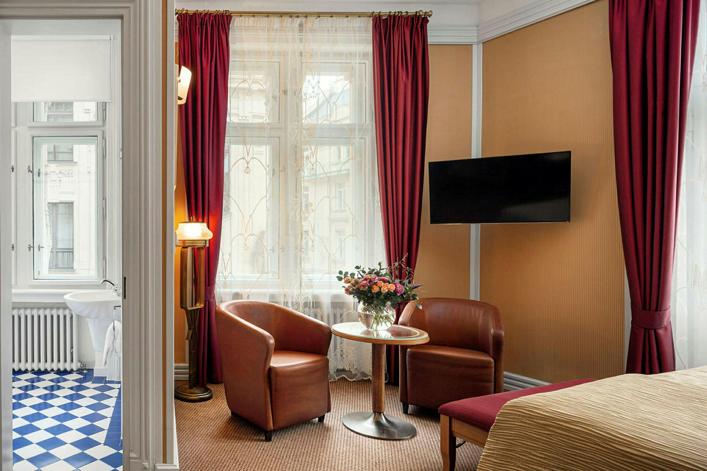Ett hotellrum med två fåtöljer och ett bord framför ett stort fönster med röda gardiner. En TV är monterad på väggen. (AI-genererad text)