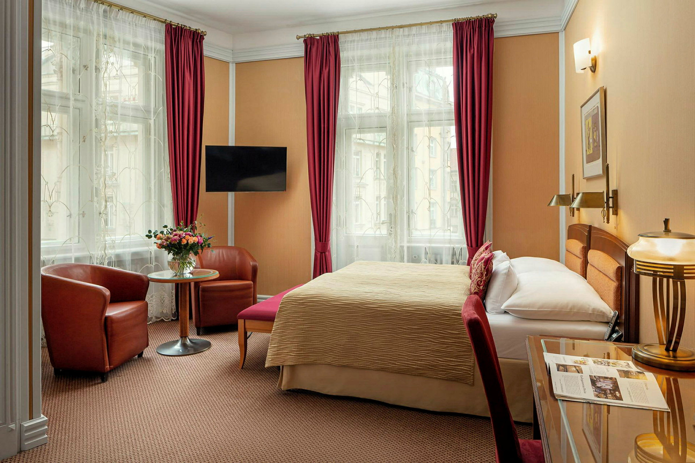 Hotellrum med en dubbelsäng, två fåtöljer vid ett bord, en TV på väggen och stora fönster med gardiner. (AI-genererad text)