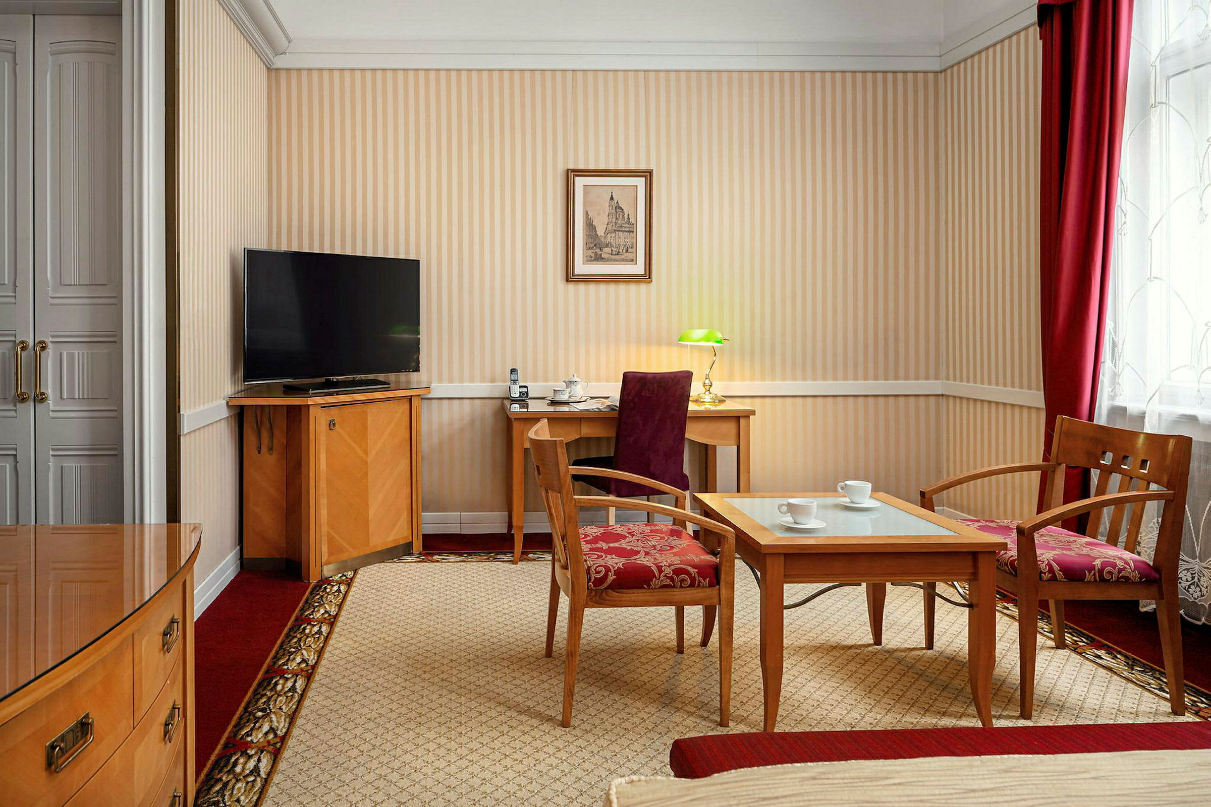 Ett hotellrum med möbler inklusive två fåtöljer, ett soffbord med glas, en skrivbordsmöbel med stol och lampa, samt en TV på en möbel. Väggarna är randiga och rummet har en röd och beige färgsättning. (AI-genererad text)