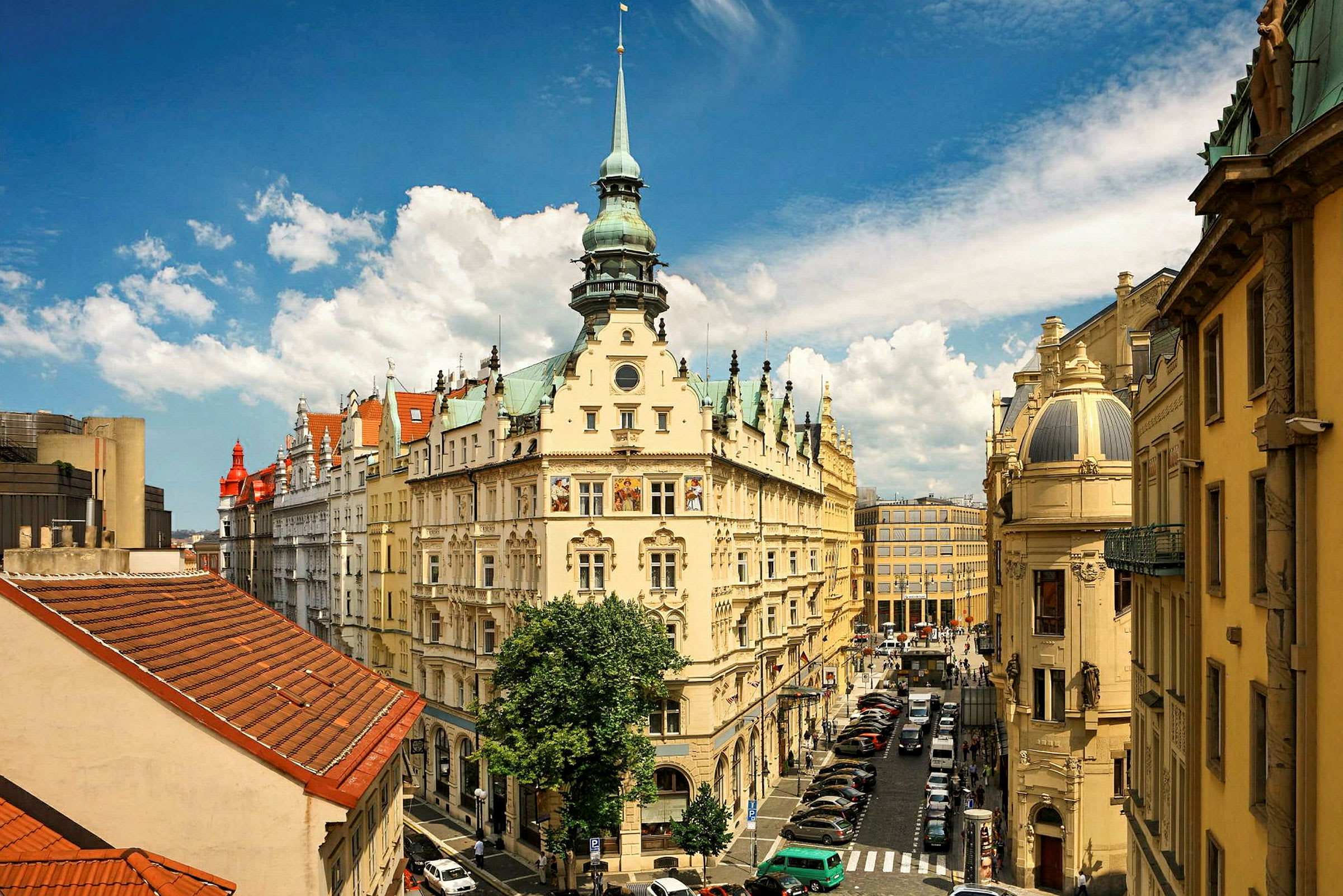 Ett europeiskt stadsmotiv med en dekorativ byggnad med torn, omgiven av fler historiska byggnader. Himlen är blå med vita moln. (AI-genererad text)