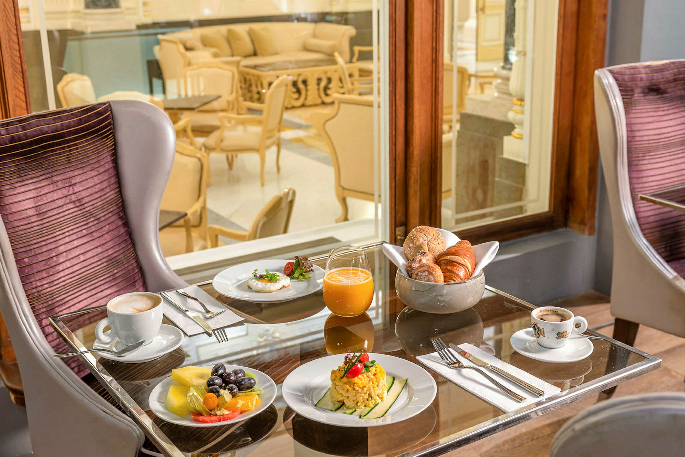 Ett dukat frukostbord med diverse mat såsom frukt, små rätter, bröd, juice och kaffe, beläget inomhus bredvid stora fönster. (AI-genererad text)