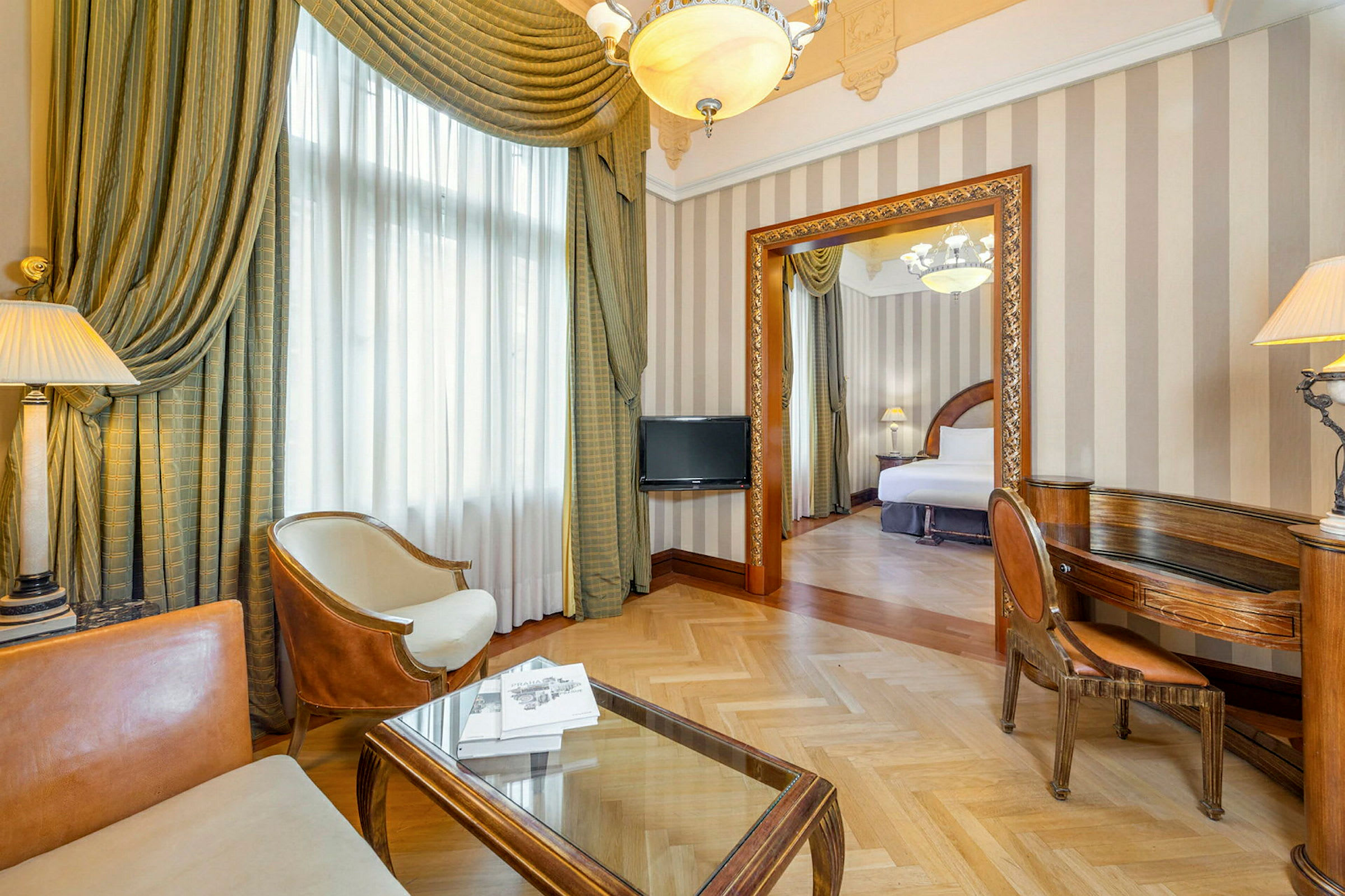 Ett elegant hotellrum med en sittgrupp bestående av stolar och bord i förgrunden, samt en öppen dörr till sovrummet med en bred säng. (AI-genererad text)