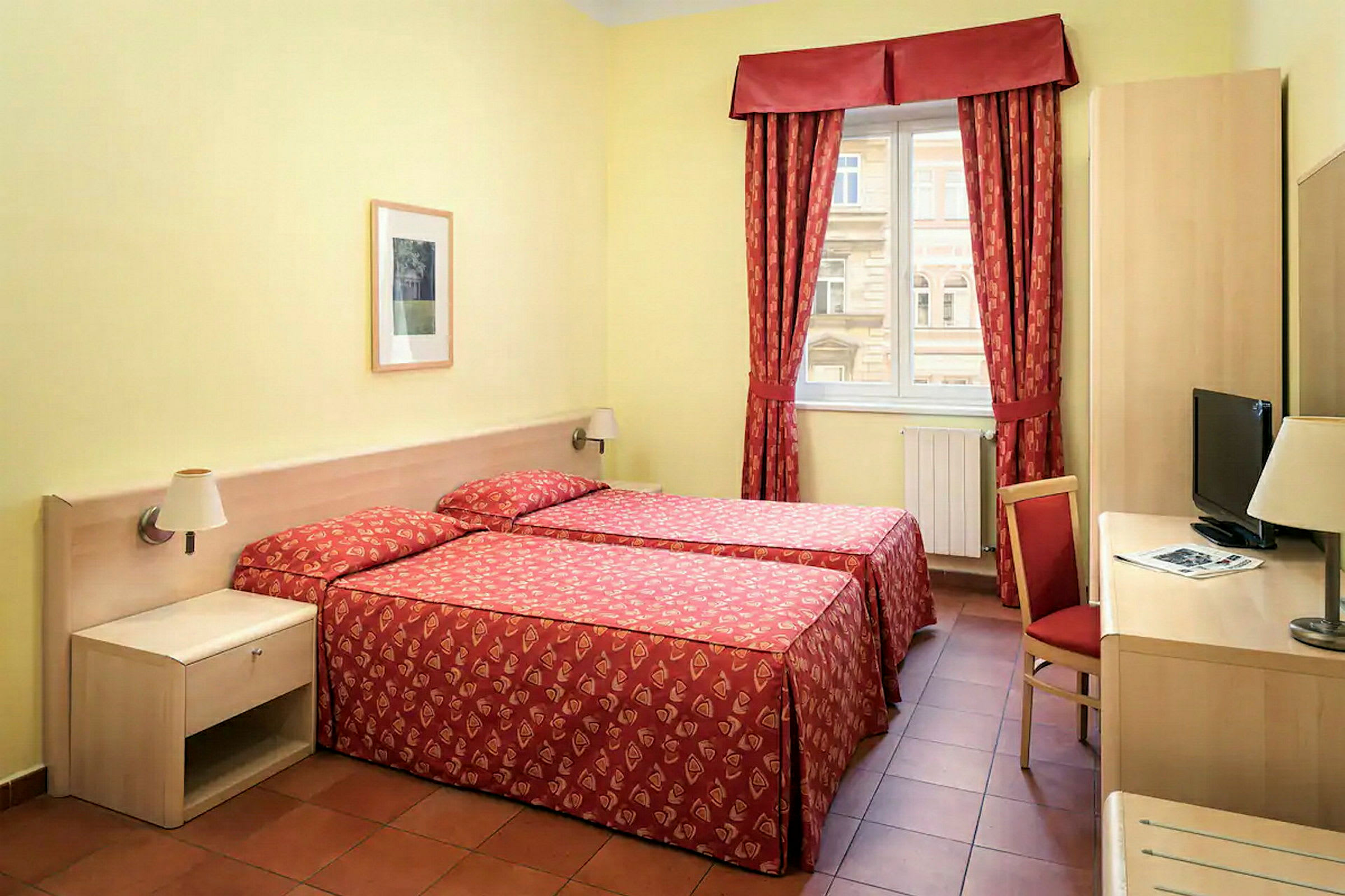 Ett hotellrum med två enkelsängar täckta med röda överkast. Det finns ett fönster med röda gardiner, ett skrivbord med en stol och en TV. (AI-genererad text)