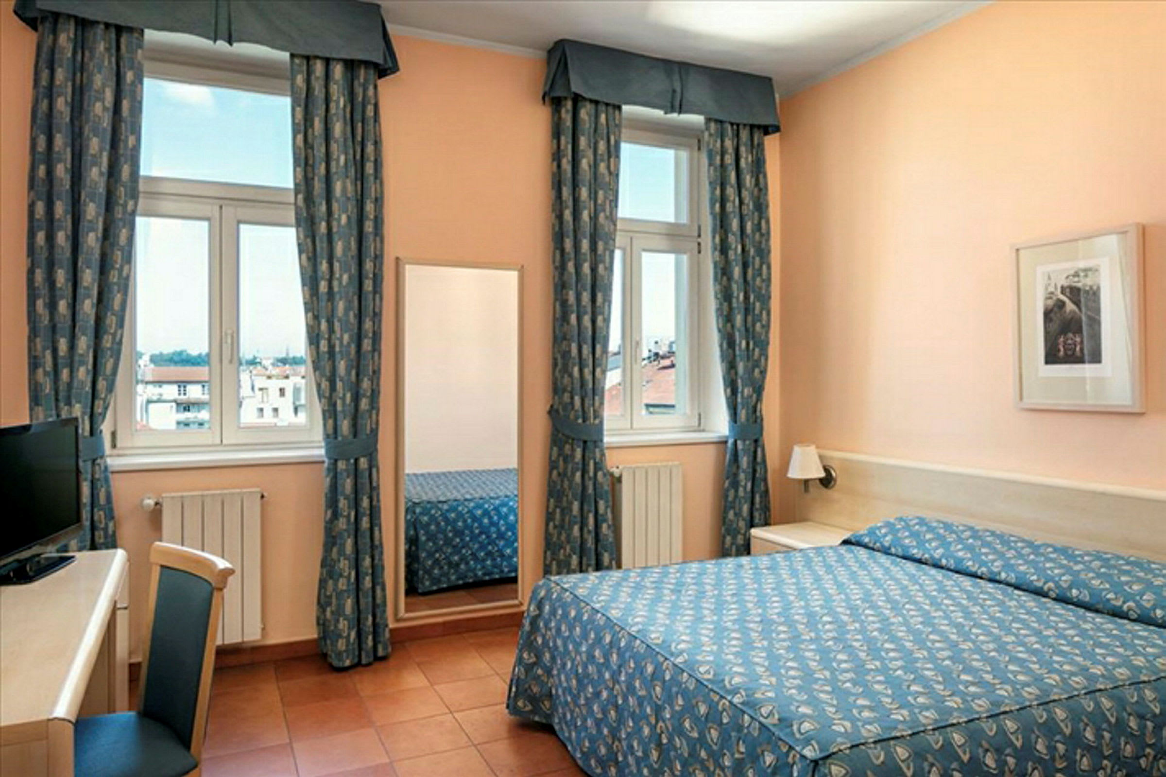 Ett hotellrum med en dubbelsäng och ett skrivbord. Fönstren är dekorerade med blå mönstrade gardiner. (AI-genererad text)