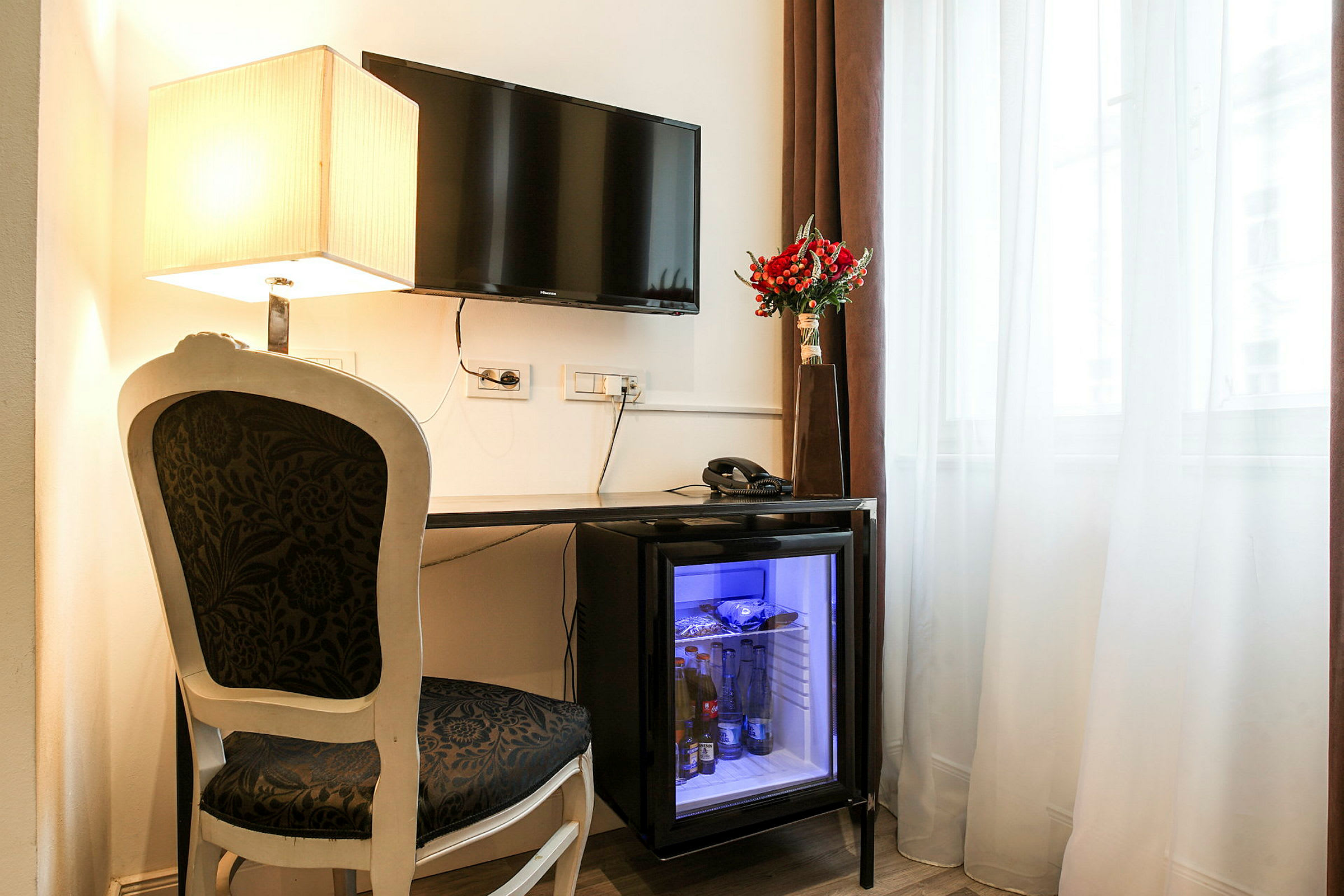 En skrivbordsplats med en stol och ett bord. Bordet har en minibar med glasdörr, en telefon och en vas med röda blommor. En fönsterplats med gardiner och en väggmonterad TV samt en bordslampa. (AI-genererad text)