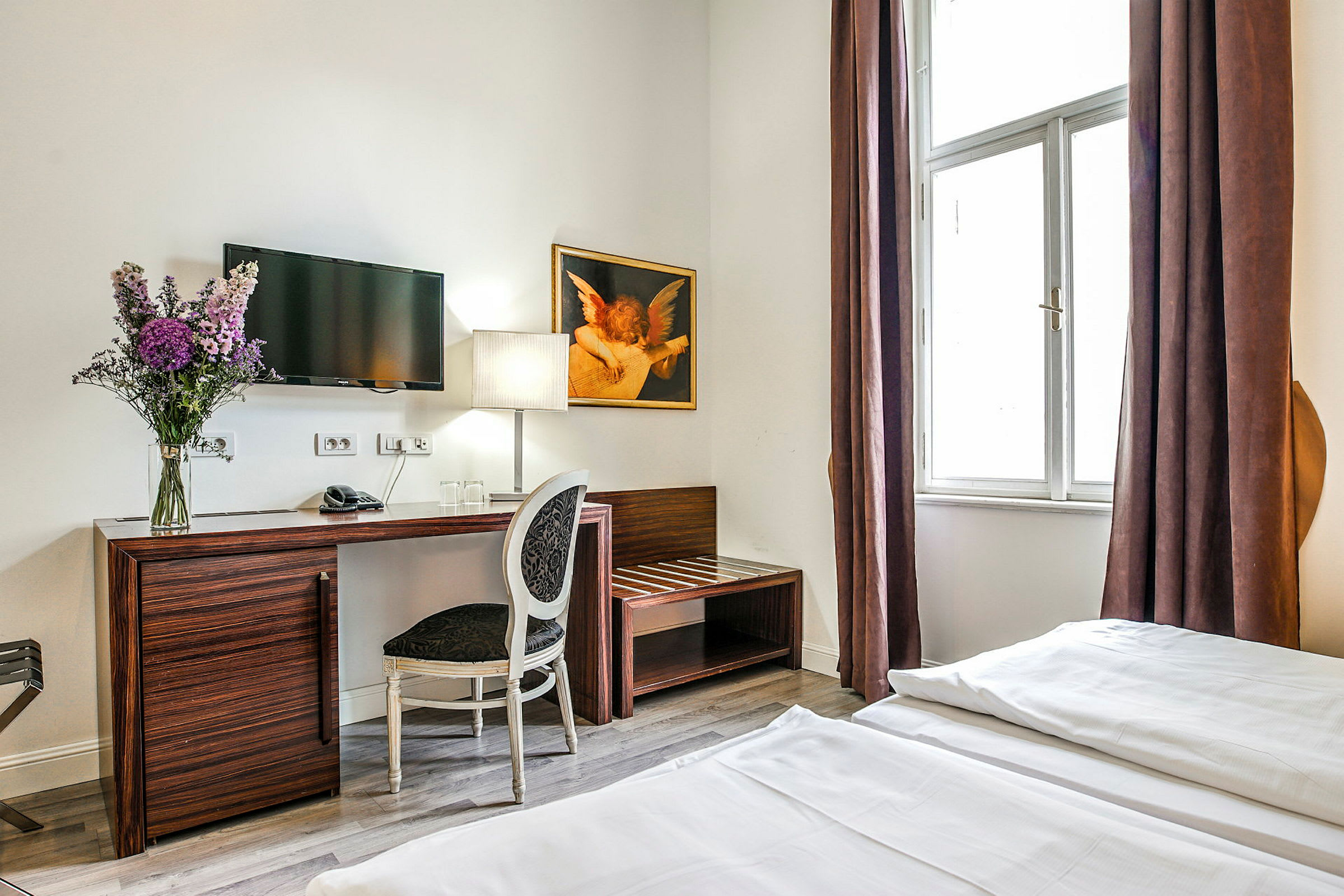 Ett hotellrum med en skrivbord och stol, en TV på väggen, en blomvas och en lampa blev placerade på bordet, samt en stor fönster med bruna gardiner. (AI-genererad text)