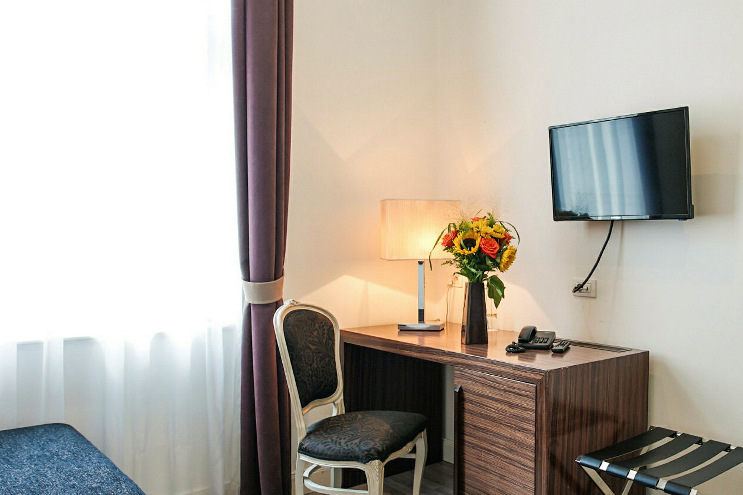 Ett skrivbord med en lampa och en blomvas placerad ovanpå, bredvid en stol och en väggmonterad TV. (AI-genererad text)