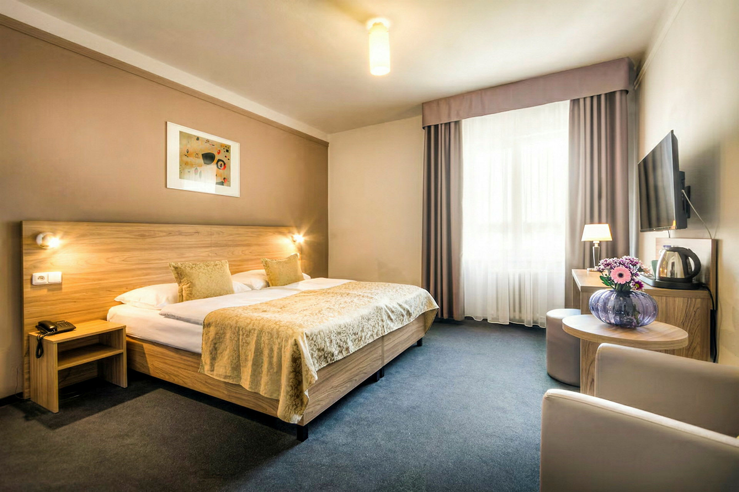 En hotellrum med en dubbelsäng, en sittgrupp, blommor på ett bord och en väggmonterad TV. (AI-genererad text)