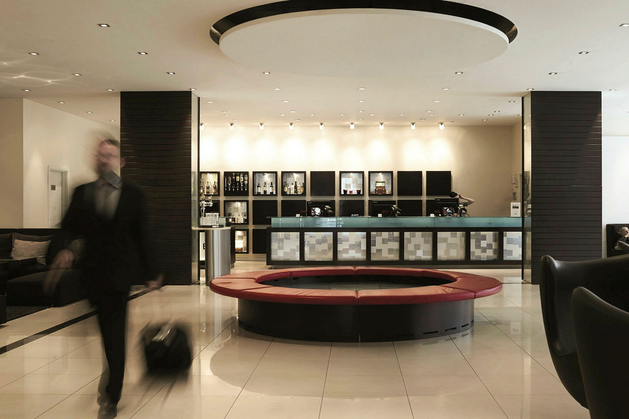 En välbelyst hotellobby med en reception, moderna sittplatser och ett runt soffområde. En person med bagage är i rörelse. (AI-genererad text)