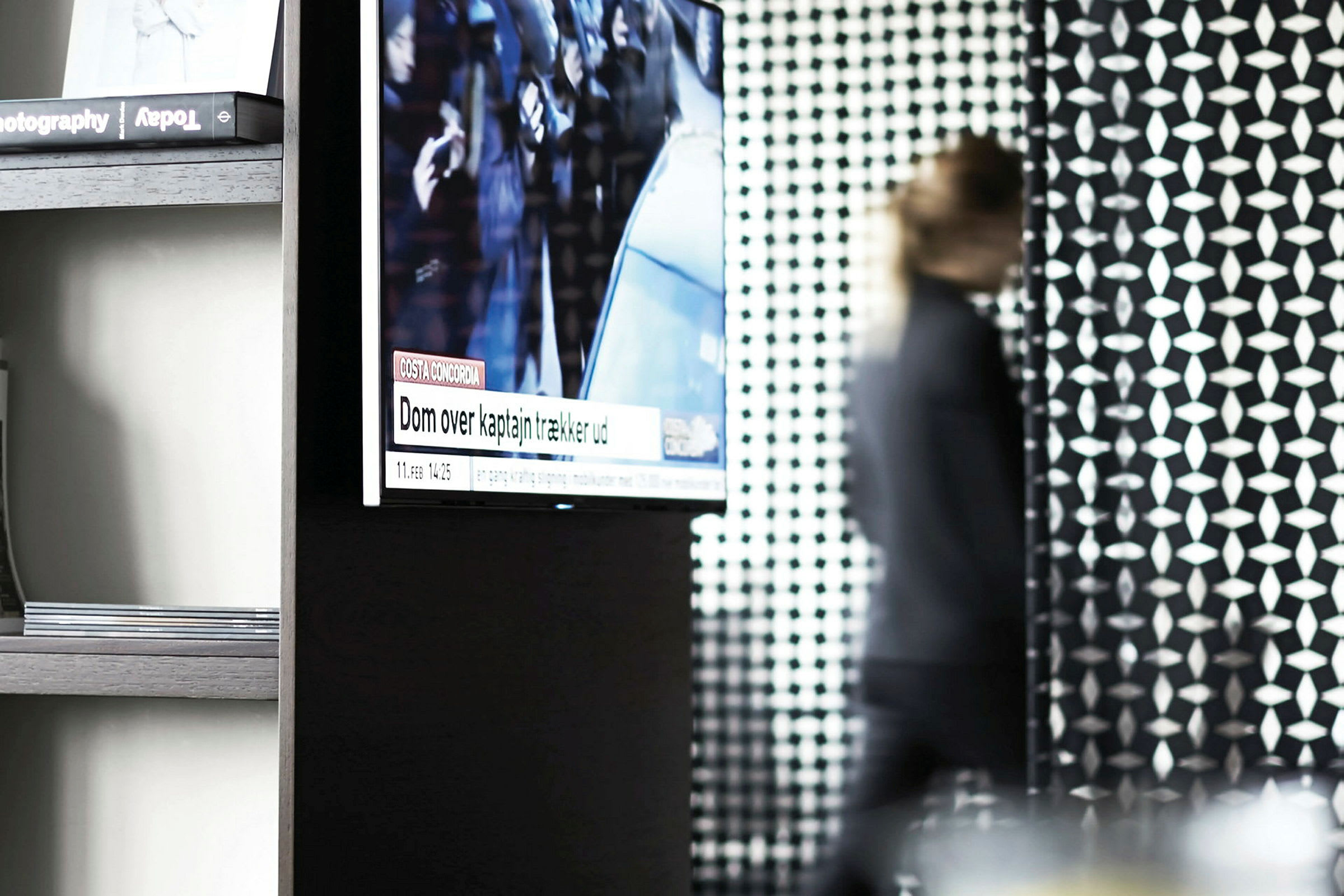 Ett rum med en väggmonterad TV som visar nyheter, en man passerar i bakgrunden med oskärpa. (AI-genererad text)