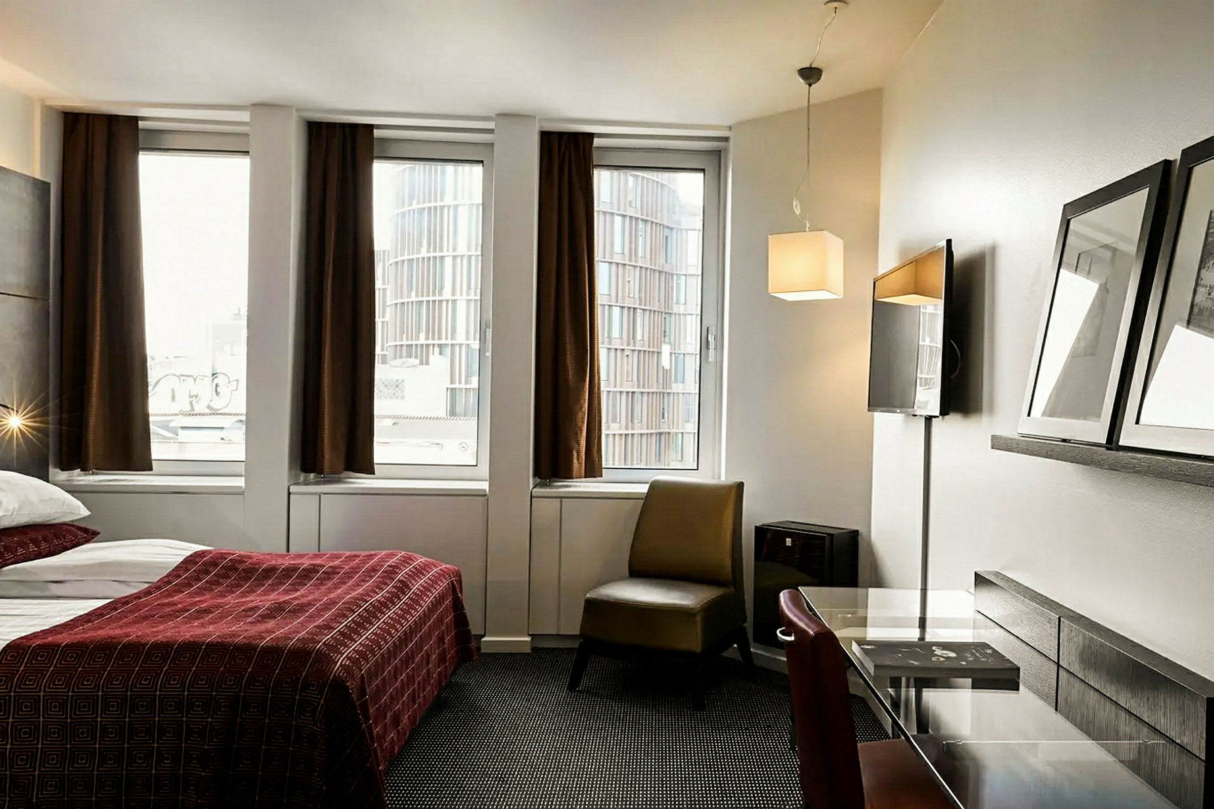 Ett hotellrum med en säng med röd sängklädsel, ett skrivbord och en stol framför tre fönster med bruna gardiner. (AI-genererad text)