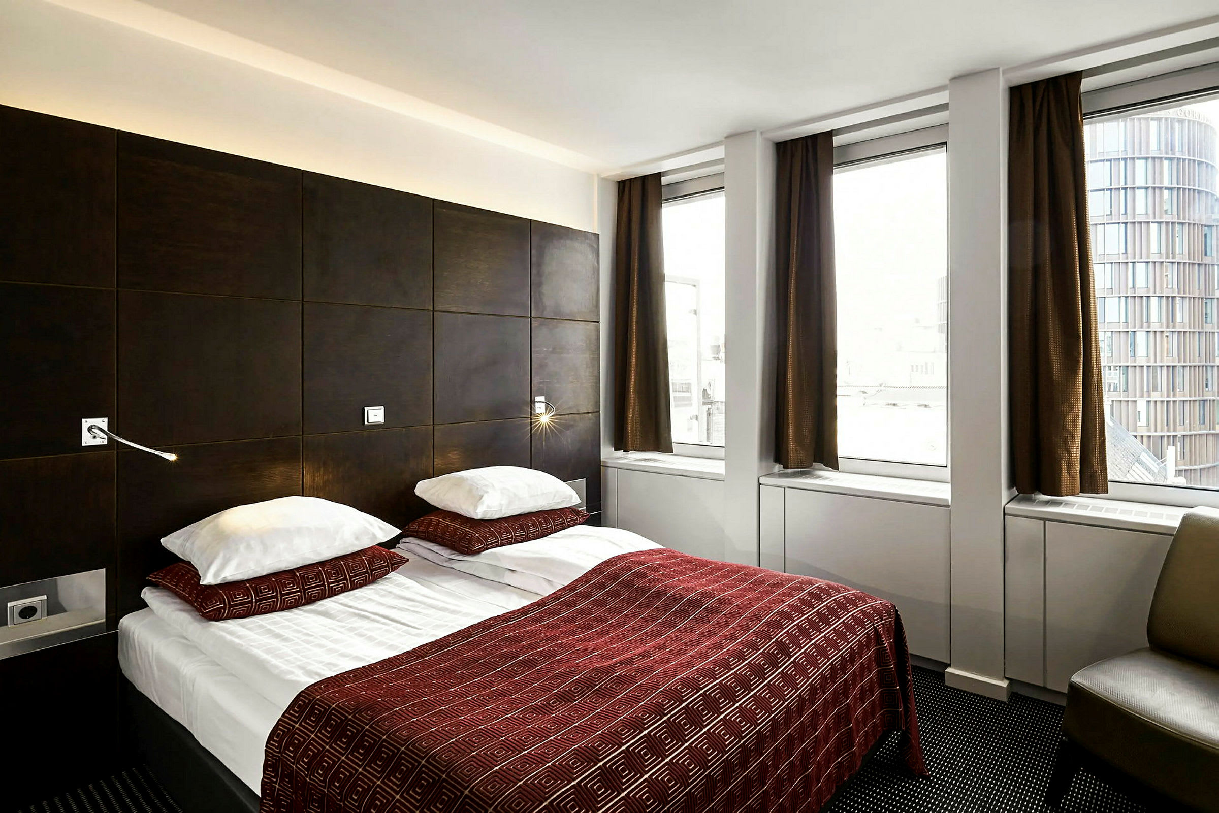Ett hotellrum med en dubbelsäng bäddad med rödmönstrade överkast och två kuddar. Rummet har mörka möbler och tre stora fönster med bruna gardiner. (AI-genererad text)