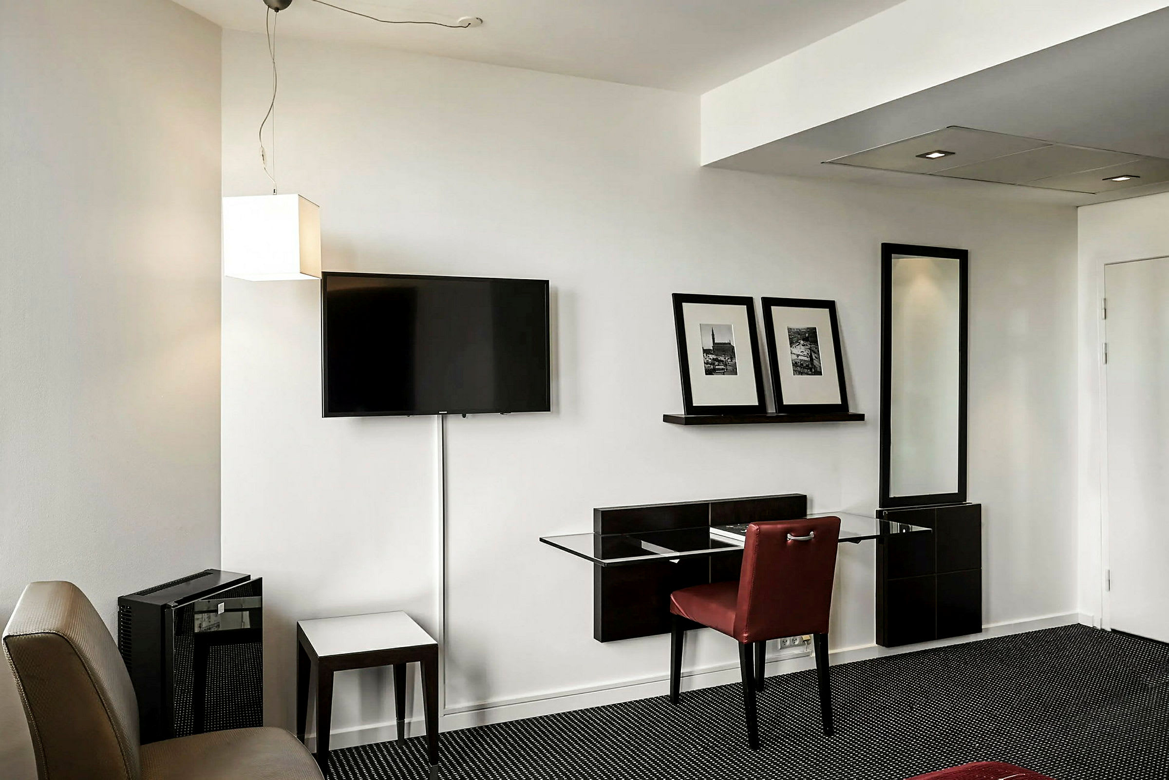 Ett hotellrum med en vägg som har en väggmonterad TV, en hylla med två inramade bilder, och en sittplats med ett bord och en stol. (AI-genererad text)