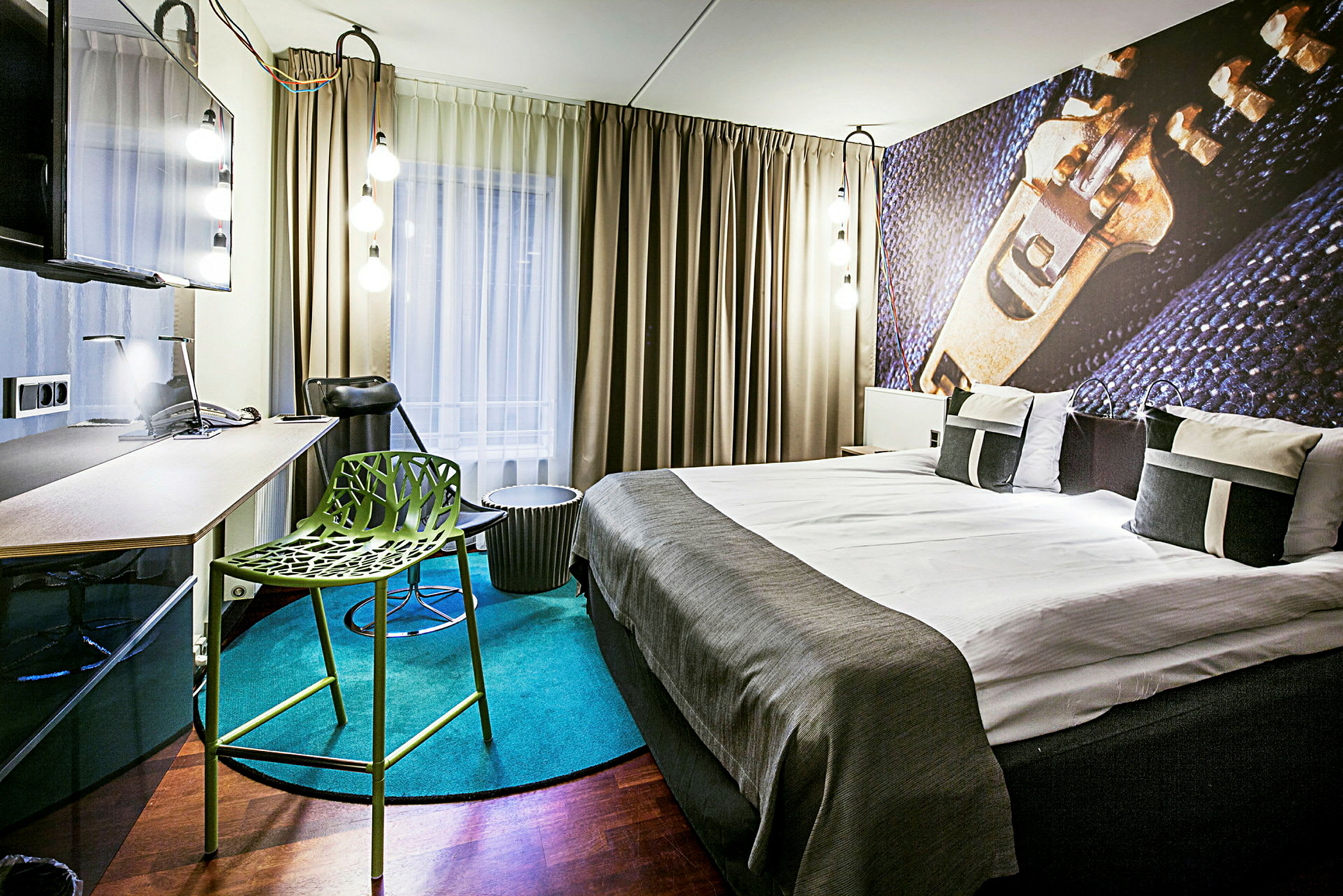 Ett modernt inrett hotellrum med en dubbelsäng, skrivbord, grönt stolsdesign och stora gardiner framför fönstret. (AI-genererad text)
