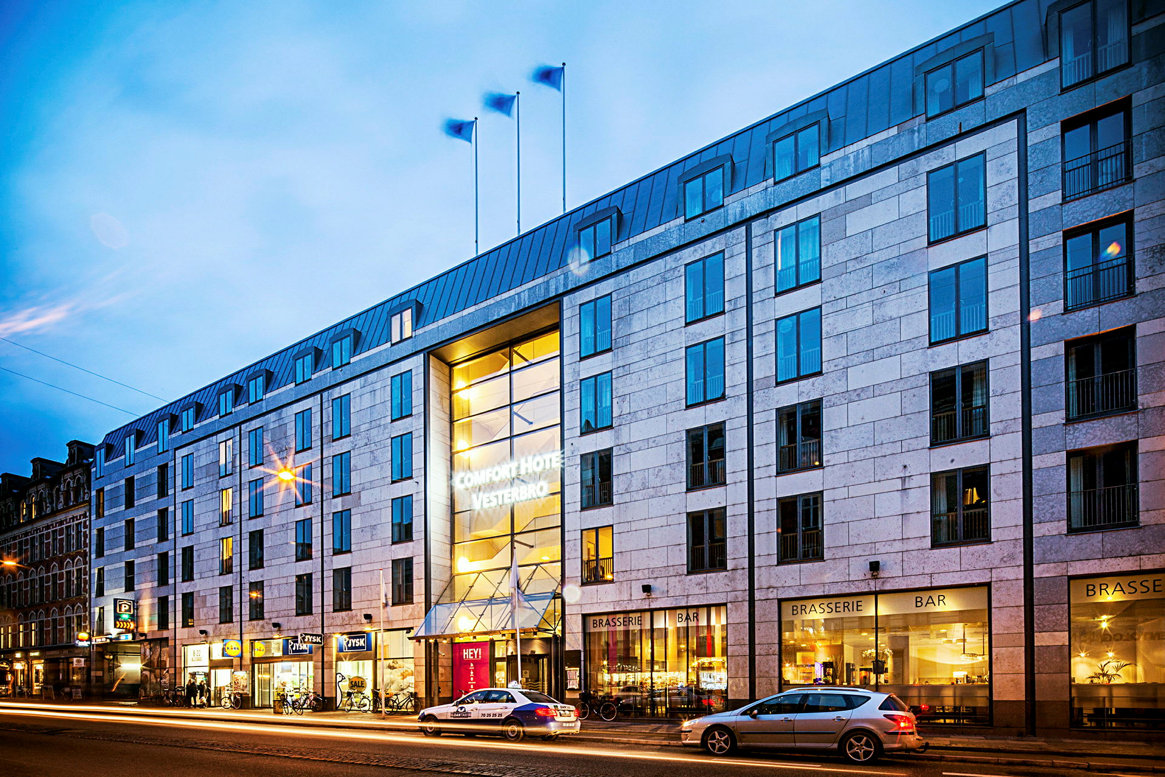 Exteriör av hotellbyggnad på kvällen, med belysning och gatan med passerande bilar. (AI-genererad text)