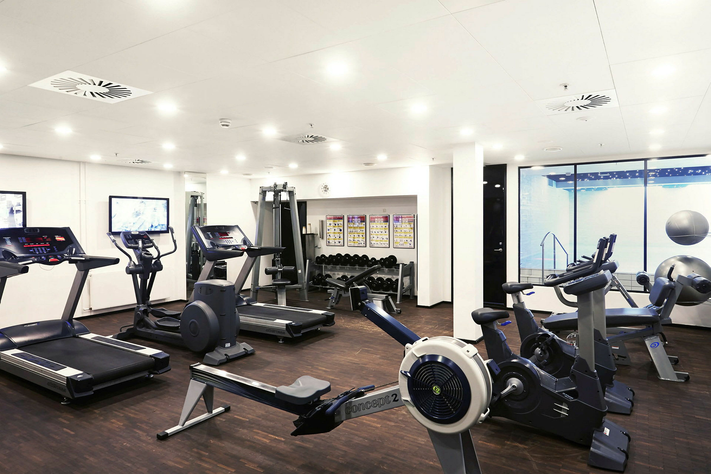 Ett gym med olika träningsmaskiner, inklusive löpband, träningscyklar och en roddmaskin. I bakgrunden syns vikter och affischer på väggen. (AI-genererad text)