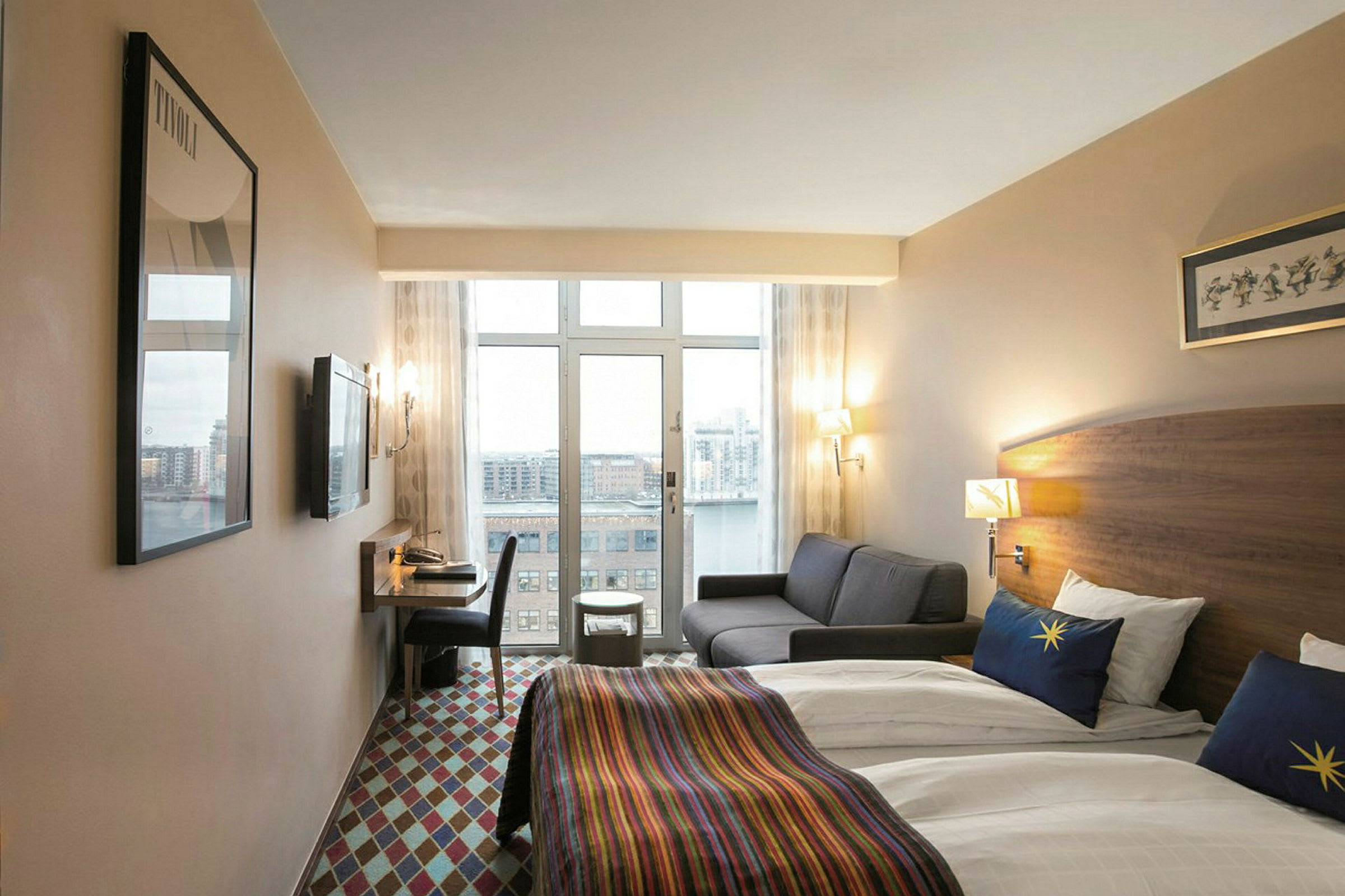 Ett hotellrum med två enkelsängar klädda i vita lakan och färgglada sängöverkast. Det finns även en grå soffa, ett skrivbord med stol och en utsikt över en stad genom stora fönster. (AI-genererad text)