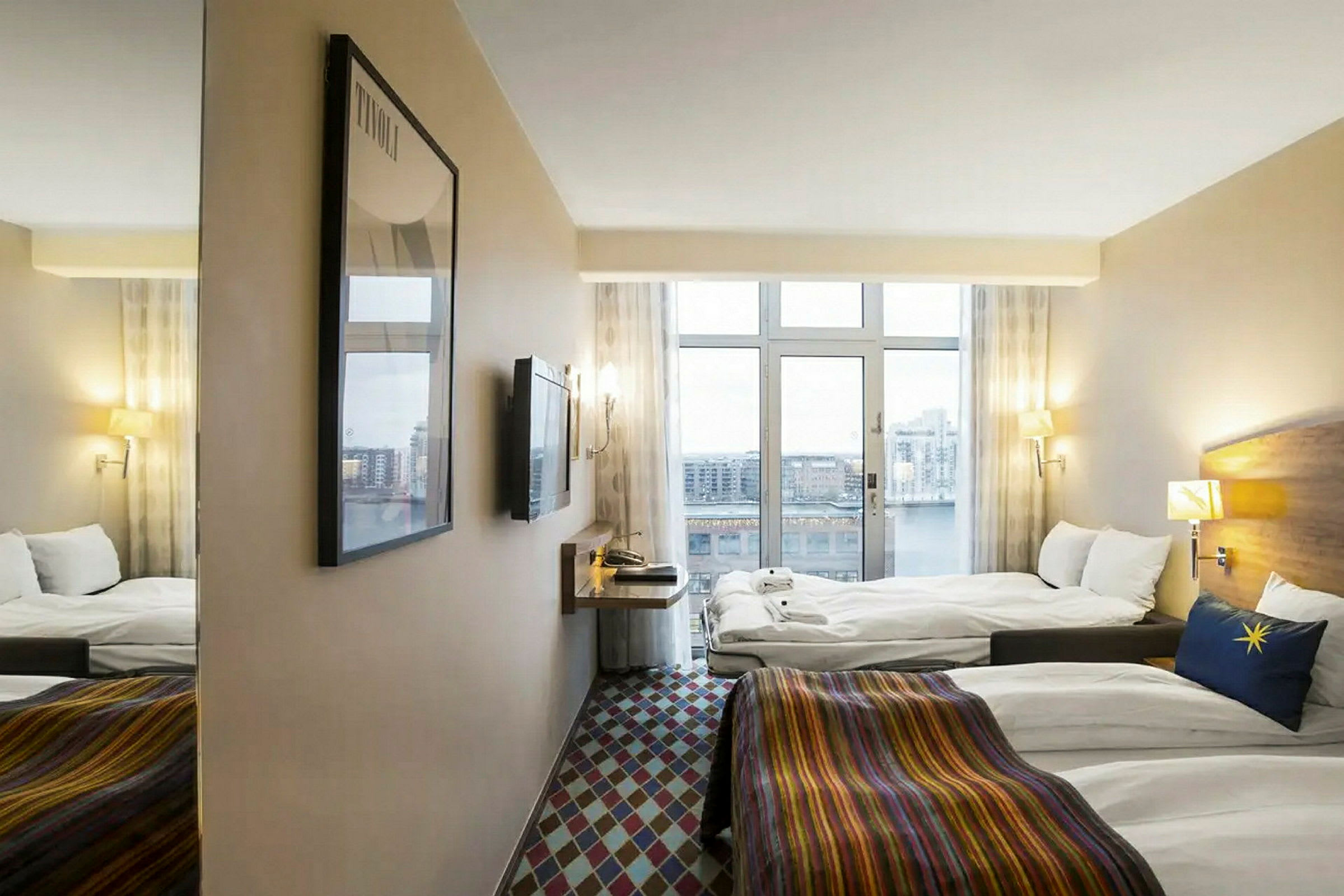 Ett hotellrum med fönster som erbjuder utsikt över en stadslandskap. Rummet är utrustat med två sängar, en väggmonterad TV och en spegel. (AI-genererad text)